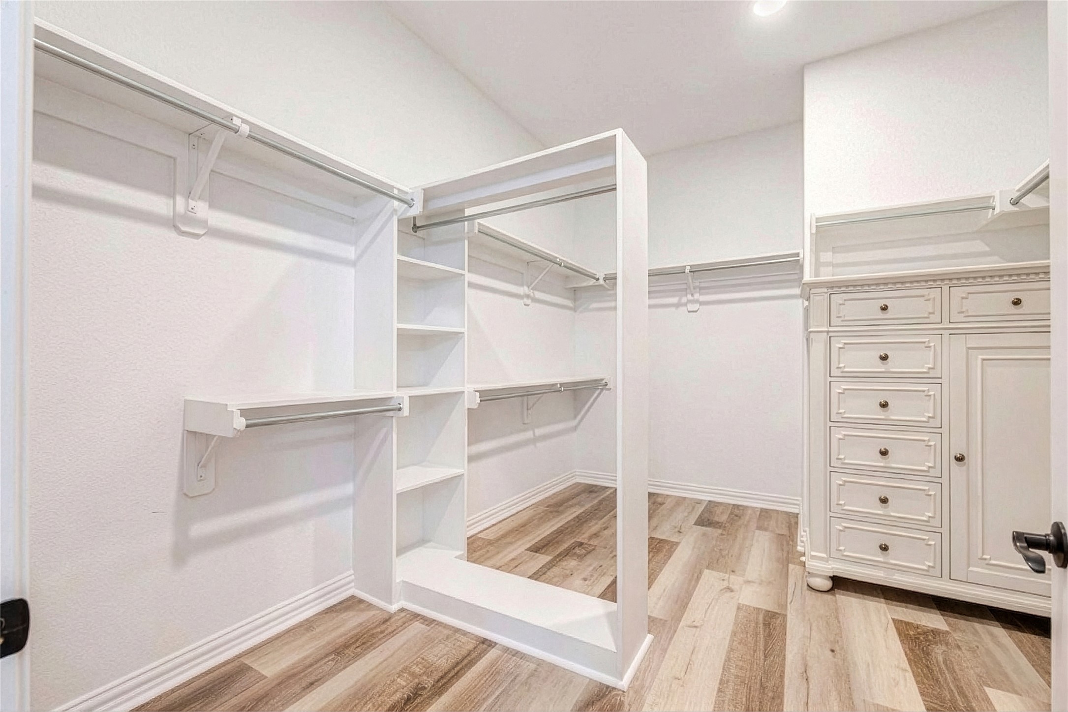 Walk-in closet with hanging rods on multiple walls, an open shelving unit, a drawer cabinet, wood-look flooring, and a ceiling light.