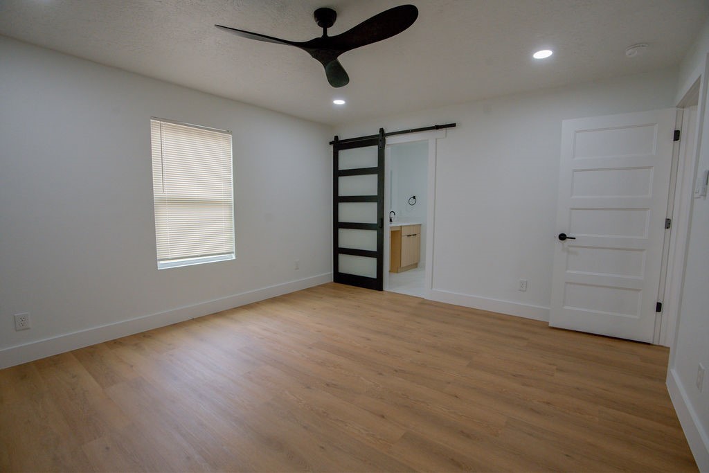 Spacious bedroom featuring sleek wood style flooring, a modern black ceiling fan, and a stylish sliding barn door leading to an en-suite bathroom.
