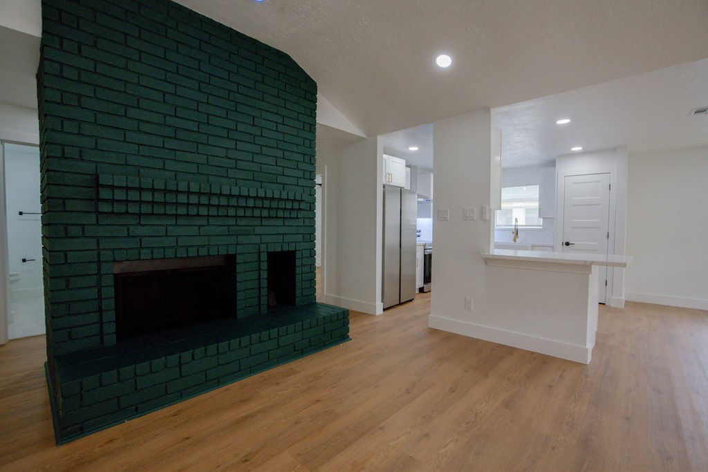 Charming living area featuring a striking dark green brick fireplace, complemented by sleek vinyl floors and recessed lighting, leading into a modern kitchen space with ample natural light.