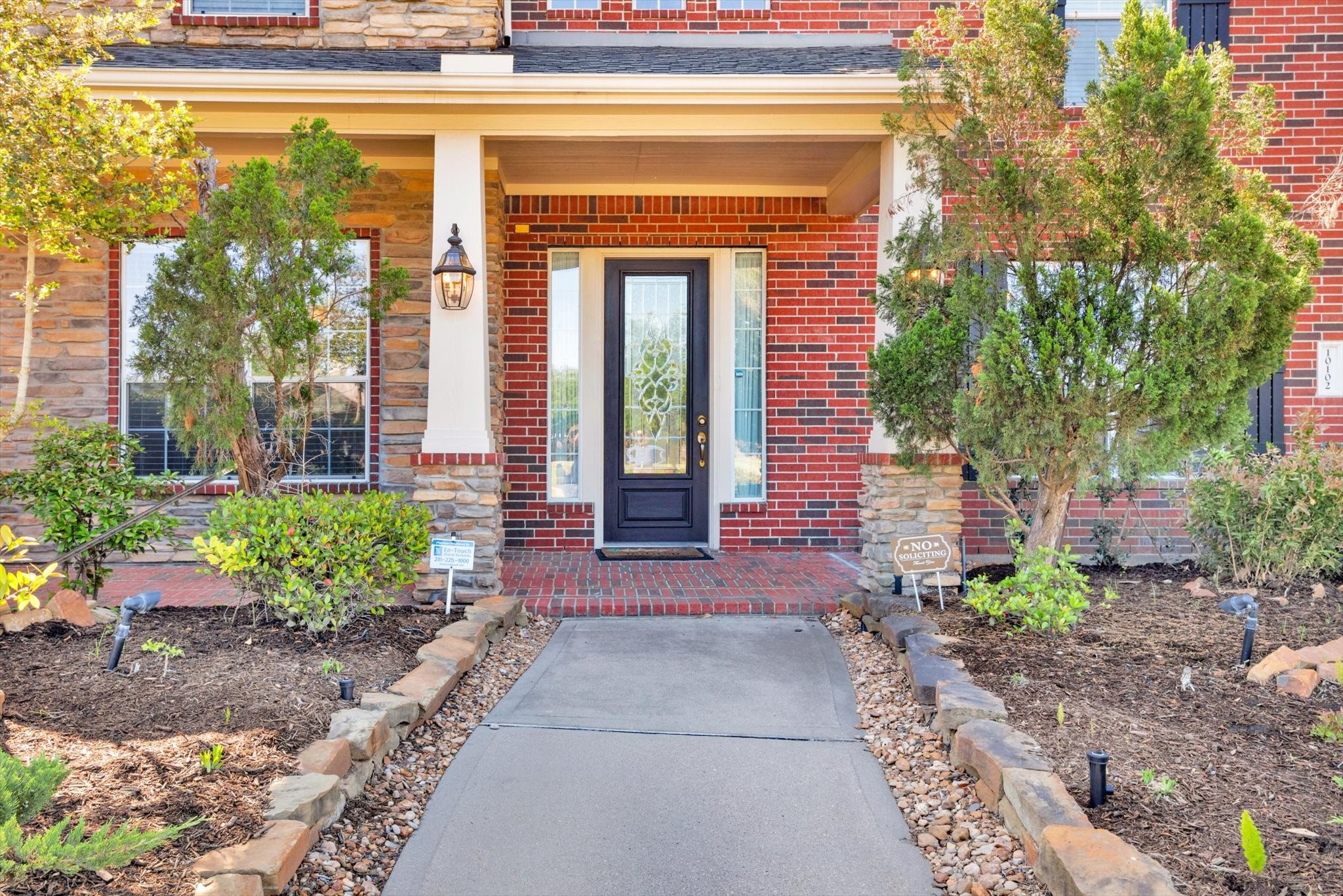 Charming brick and stone exterior with a welcoming front porch. The entryway features a stylish black door flanked by sidelights, surrounded by manicured landscaping and a stone-lined walkway. Ideal for creating a warm first impression.