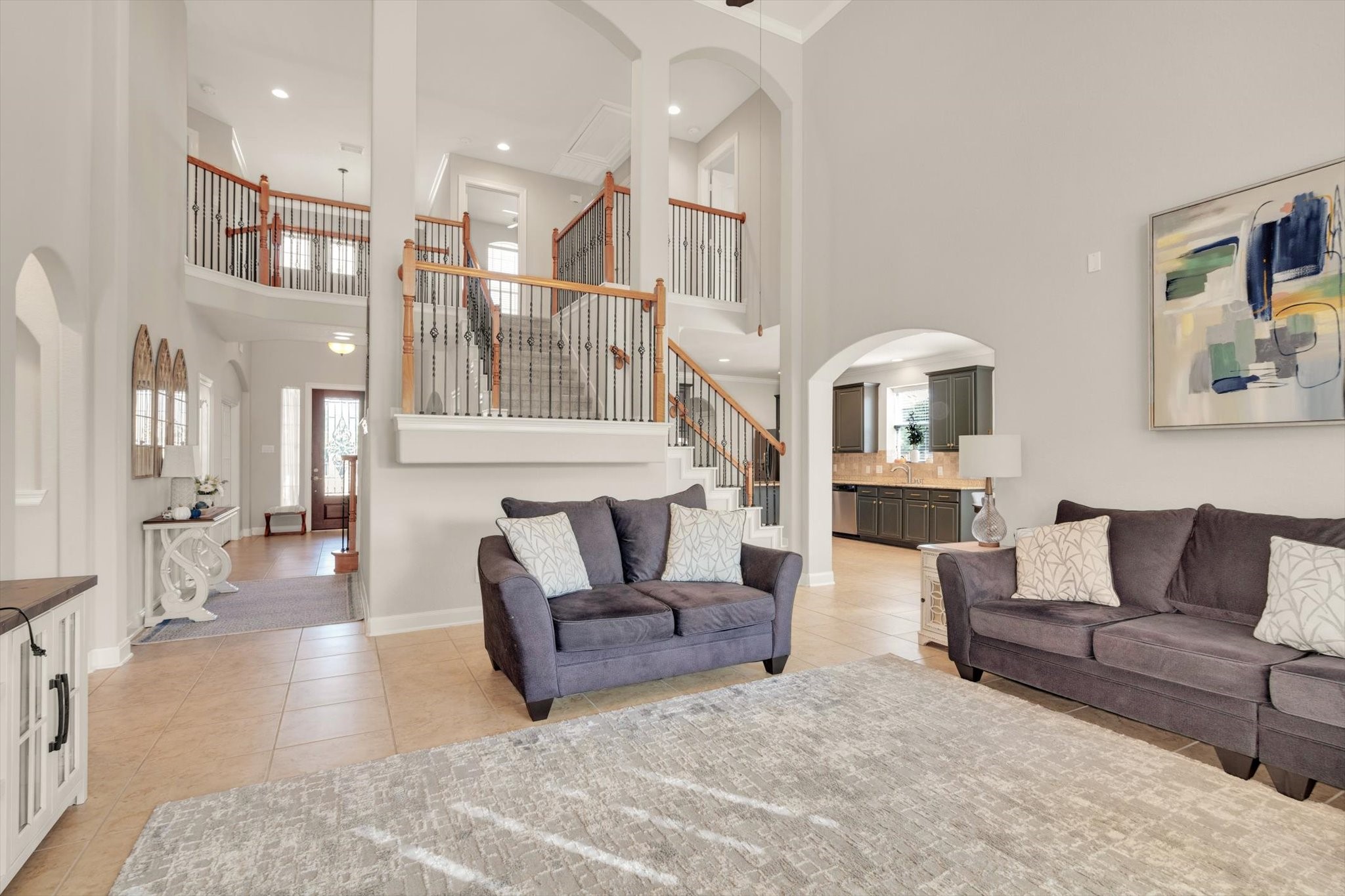 This spacious living room features high ceilings, a modern open staircase with wooden accents, and a bright, neutral color palette. It flows seamlessly into a kitchen area, creating an inviting open-concept space perfect for entertaining.