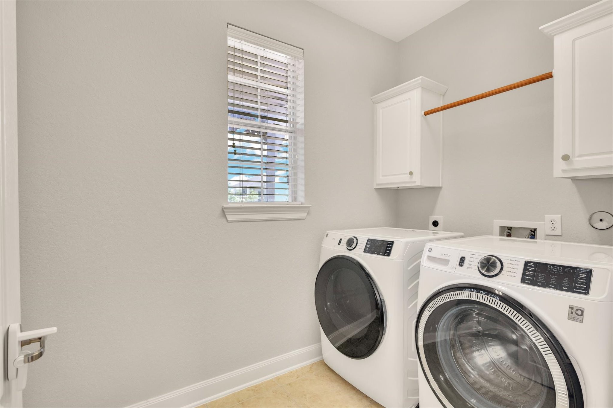This is a bright, modern laundry room featuring a washer and dryer set, white cabinetry for storage, a hanging rod, and a window for natural light.