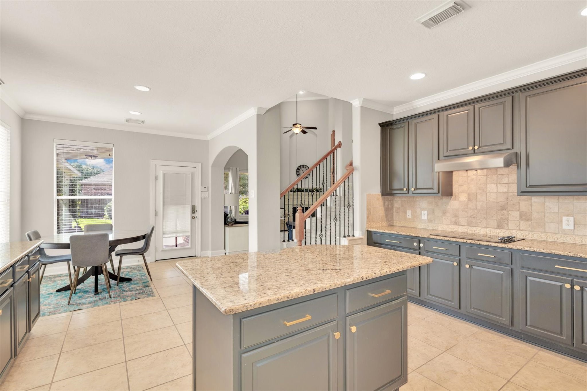 This photo showcases a spacious kitchen with elegant gray cabinets, granite countertops, and tile flooring. The room is well-lit with natural light from large windows. There's an open dining area and a view of a staircase, emphasizing a modern and inviting layout.