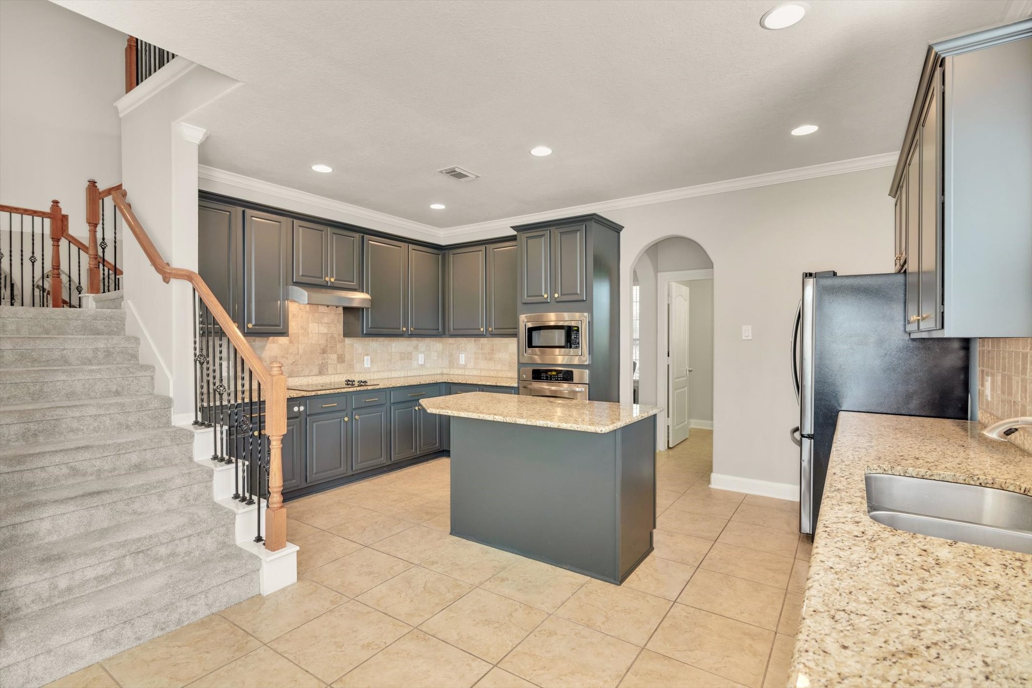 This kitchen features modern dark cabinetry, granite countertops, and stainless steel appliances. It includes a central island and an adjacent staircase with carpeted steps and wooden railings, creating an elegant and functional space.