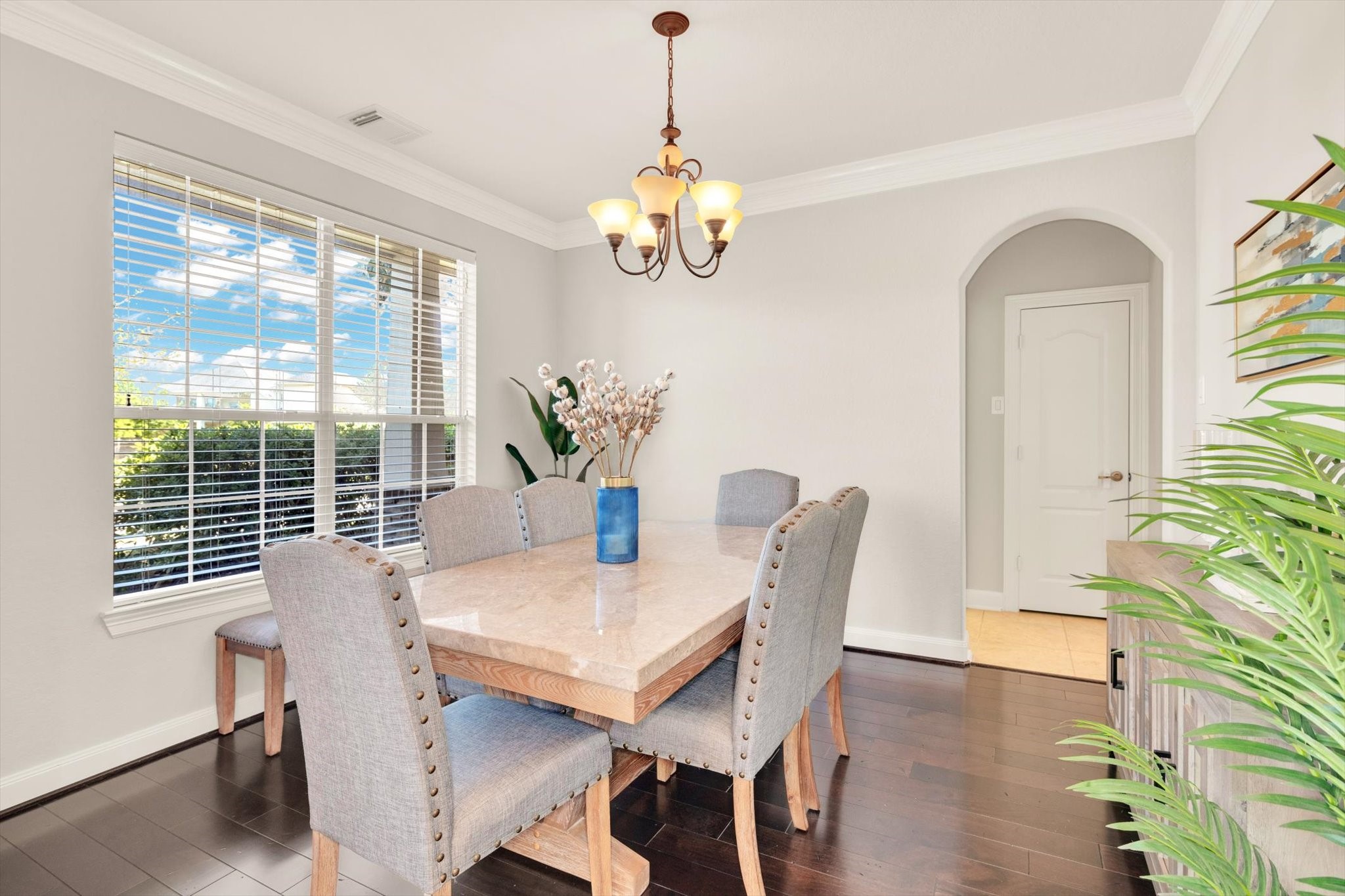 Bright and inviting dining room with a large window, elegant chandelier, and a stylish marble table with upholstered chairs. The space features dark hardwood floors and a subtle arch leading to another area, enhancing its charm.