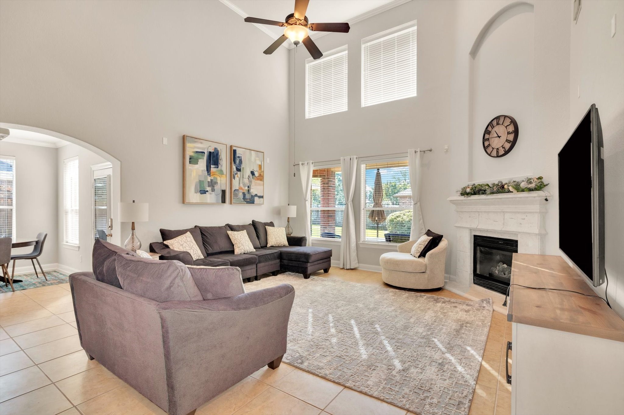 This spacious living room features a high ceiling with large windows for ample natural light. It includes a cozy fireplace, modern decor, and a comfortable sectional sofa. The open layout flows into a dining area, enhancing the home's inviting atmosphere.