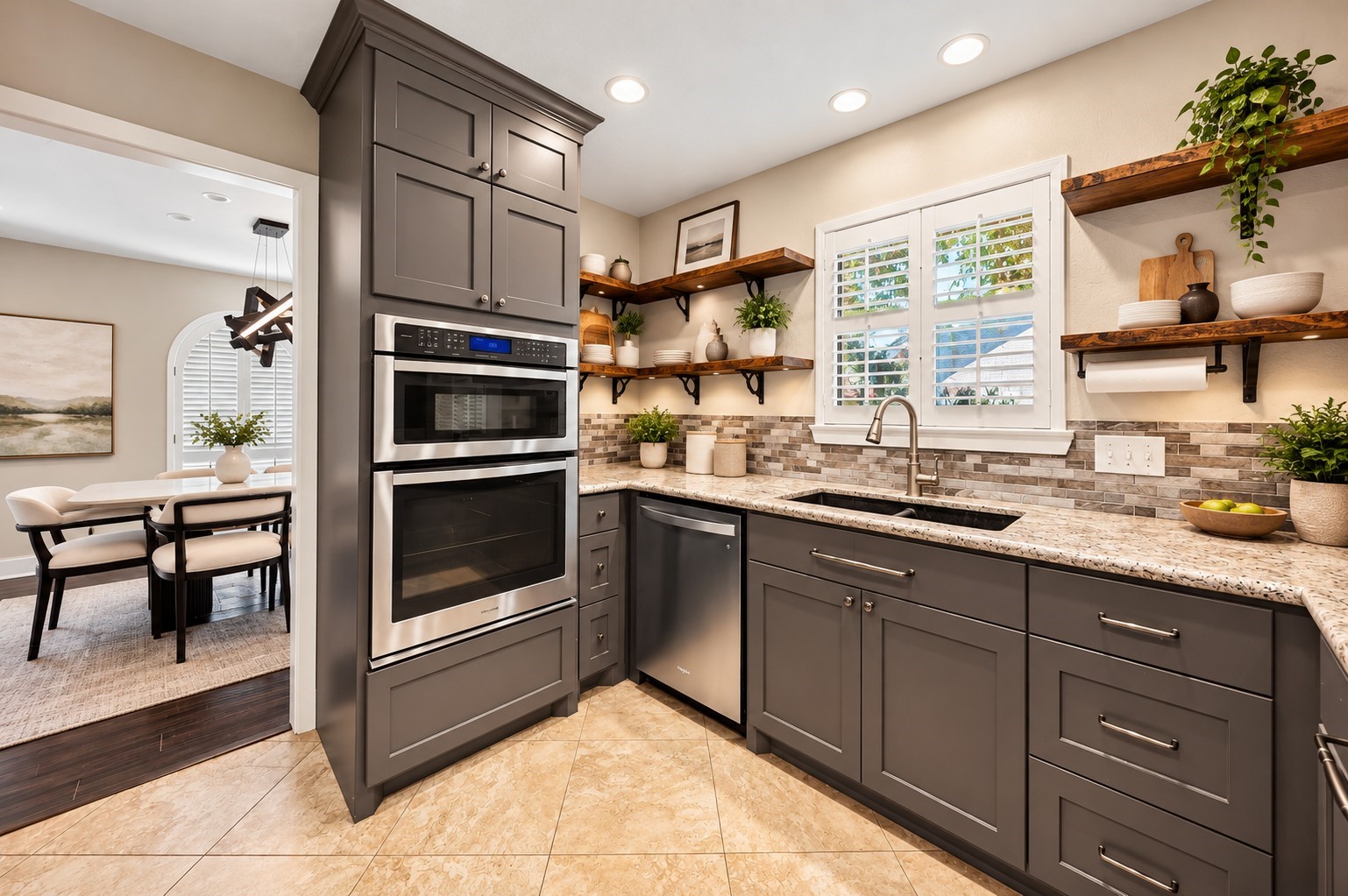 Kitchen (Virtually Staged)
Additional kitchen view highlighting stainless steel double ovens, granite countertops, gray cabinetry, decorative tile backsplash, and open wood shelving. Conveniently positioned near the formal dining room for effortless entertaining.