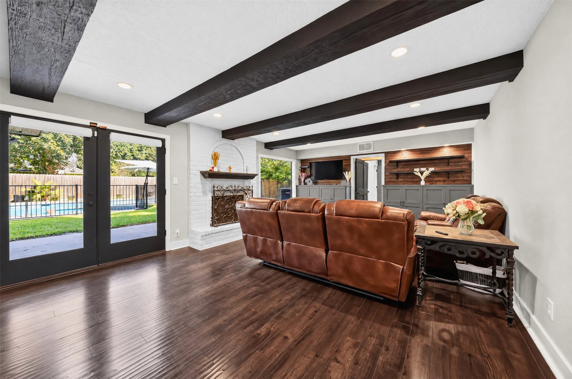 Spacious Living Room / Backyard Access
Beautifully appointed living area featuring dramatic exposed wood ceiling beams, rich wood-look flooring, a painted brick fireplace, and French doors overlooking the backyard pool. An inviting space designed for relaxation and entertaining.