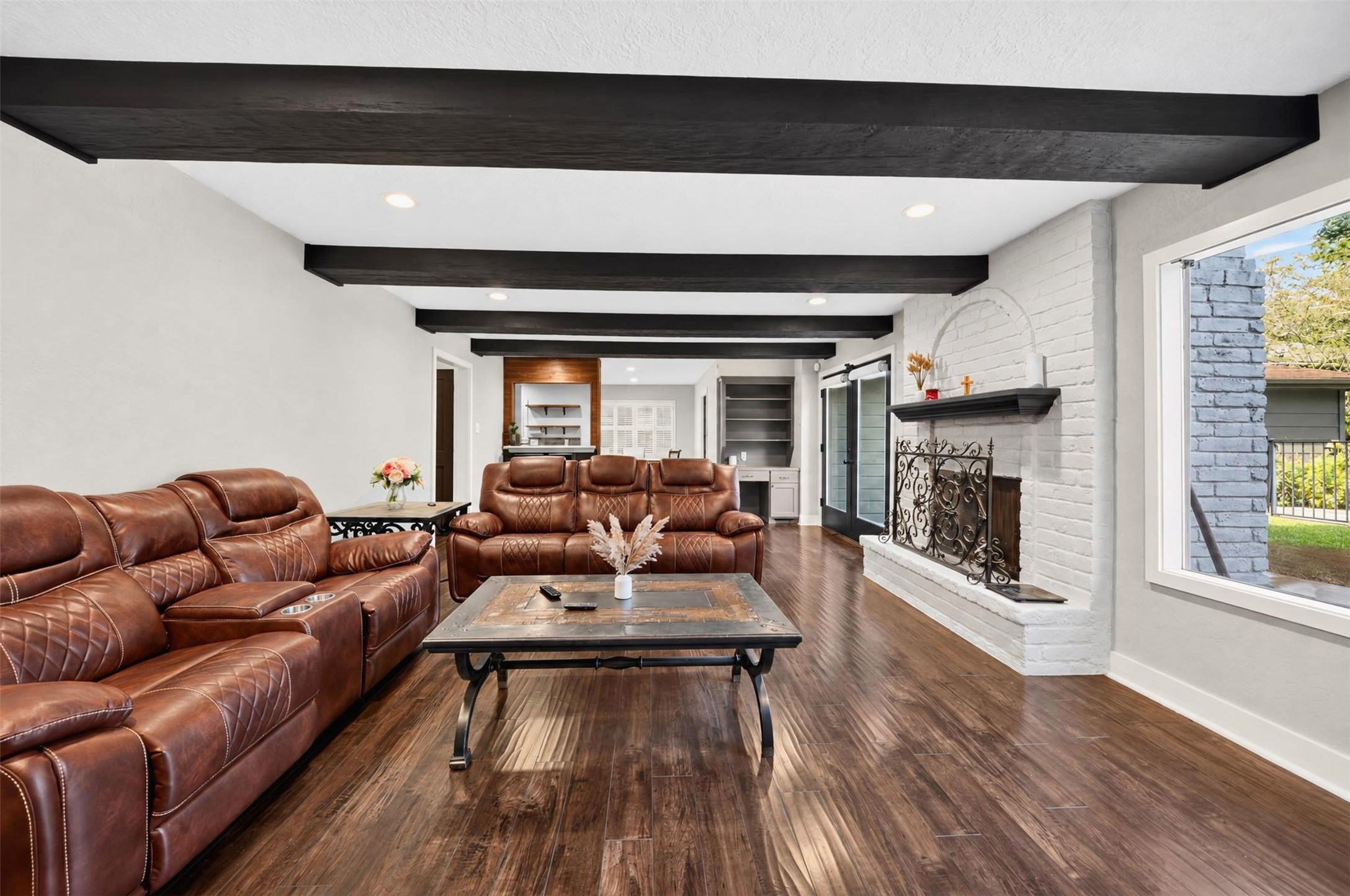 Living Room: Spacious living room featuring rich wood-look flooring, striking exposed ceiling beams, a white brick fireplace, and abundant natural light.