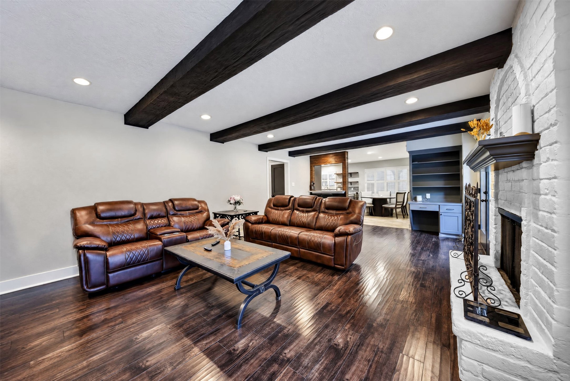 Living Room / Additional View
Expansive living room showcasing striking exposed wood beams, rich wood-look flooring, and a beautiful white brick fireplace. The open design creates an inviting atmosphere with seamless access to the adjoining breakfast area.