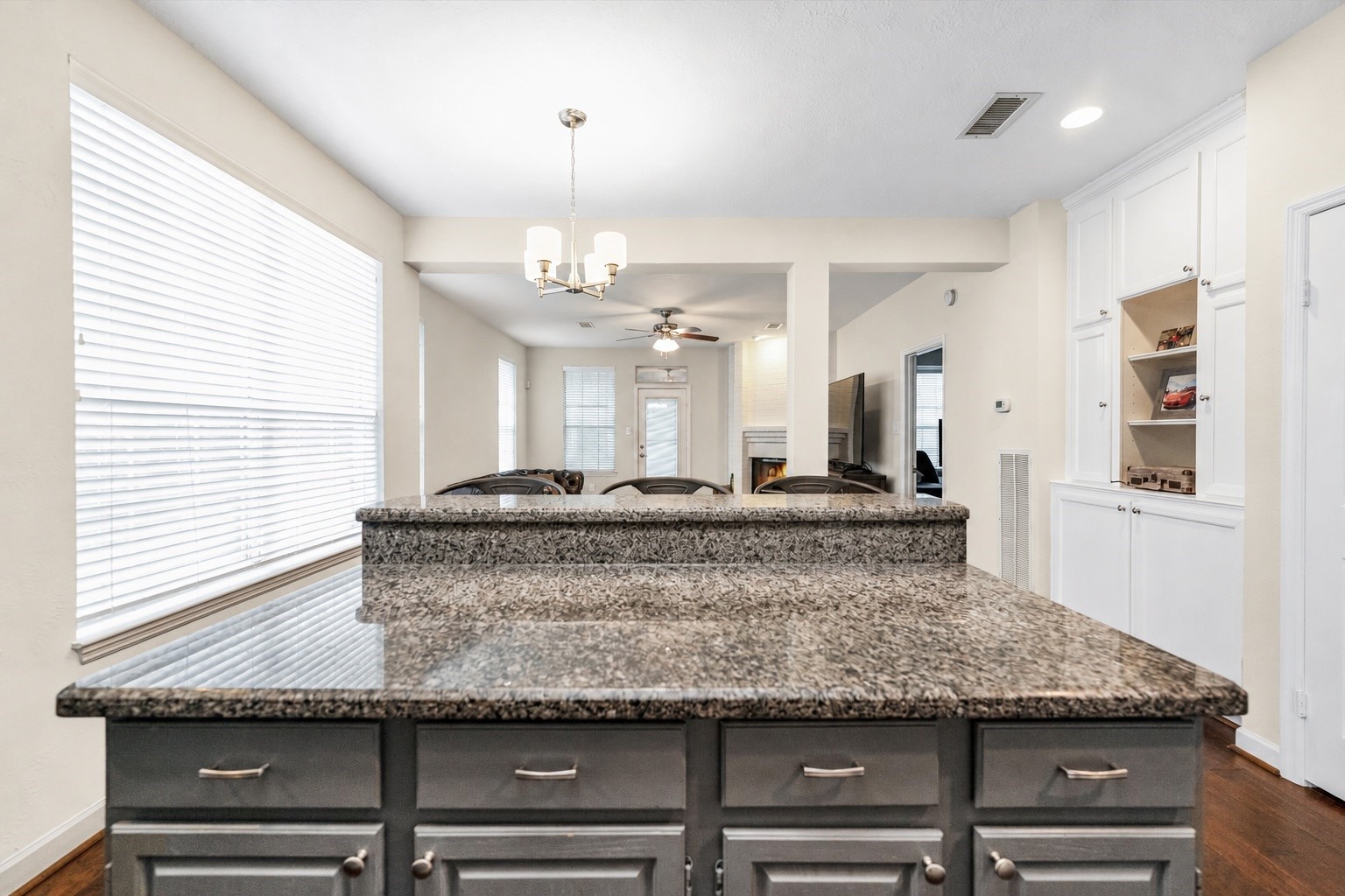Spacious kitchen featuring a sleek granite island, ample cabinetry, and a view into the bright, open-concept living area with built-in shelving and a cozy fireplace. Spacious kitchen featuring a sleek granite island, ample cabinetry, and a view into the bright, open-concept living area with built-in shelving and a cozy fireplace.