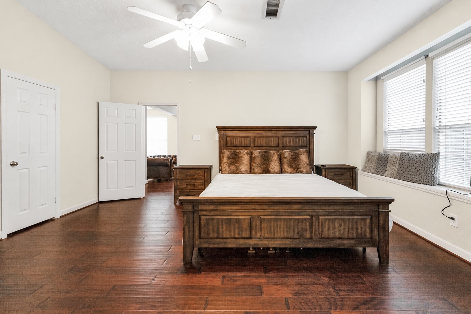 Spacious main bedroom with elegant hardwood floors, featuring a large wooden bed, ample natural light from multiple windows, and a charming window seat for relaxation.