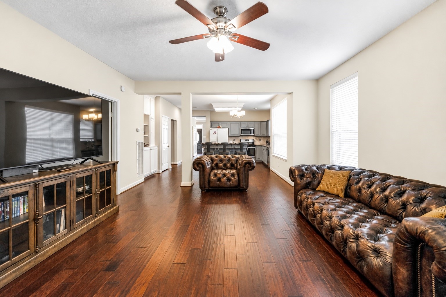 Spacious living area with rich hardwood floors, elegant leather seating, and a ceiling fan, seamlessly flowing into a modern kitchen with stainless steel appliances. Spacious living area with rich hardwood floors, elegant leather seating, and a ceiling fan, seamlessly flowing into a modern kitchen with stainless steel appliances.