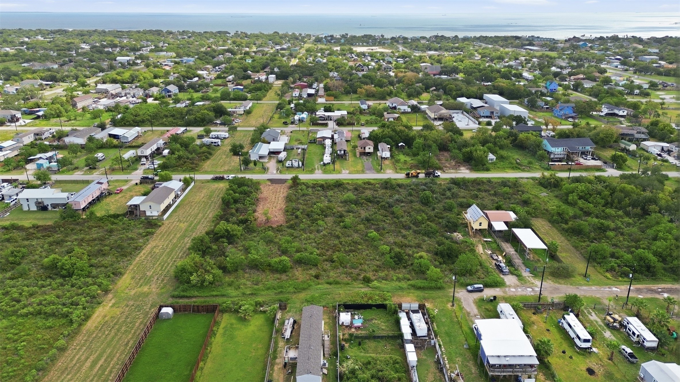 Aerial view from above the road looking across the property toward Galveston Bay. A cleared pad with a gravel driveway apron sits at the road frontage. Established neighborhood homes line the street across the way.