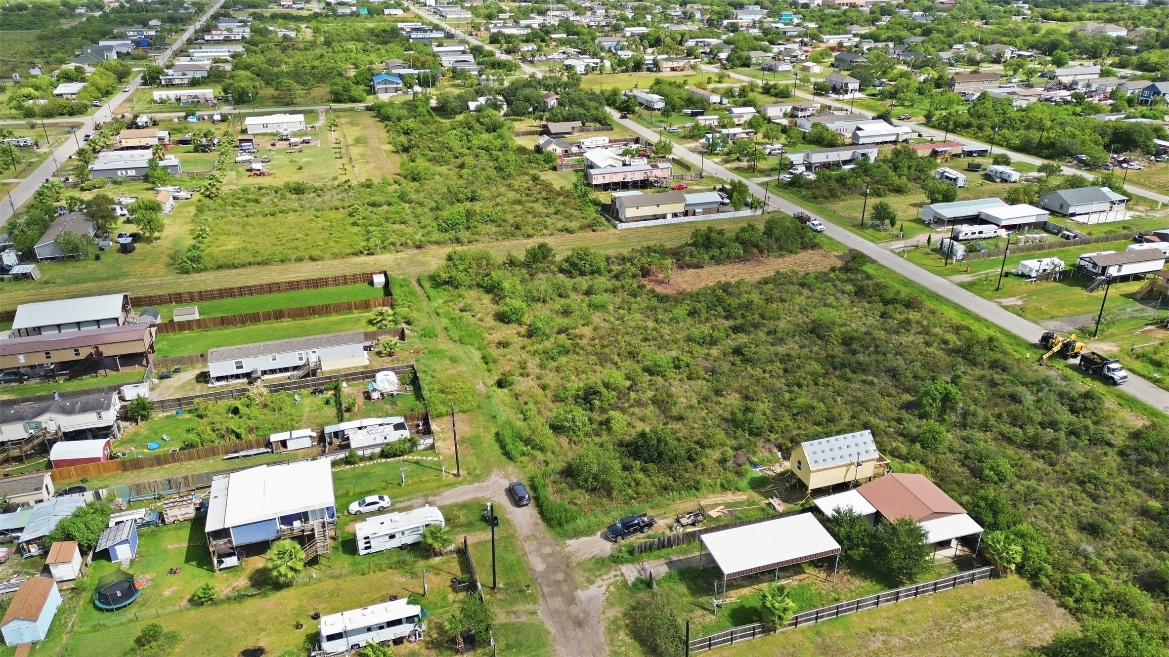 Aerial from the rear of the property looking out over the neighborhood. A gravel access road runs along one side, and the paved street is at the far edge.