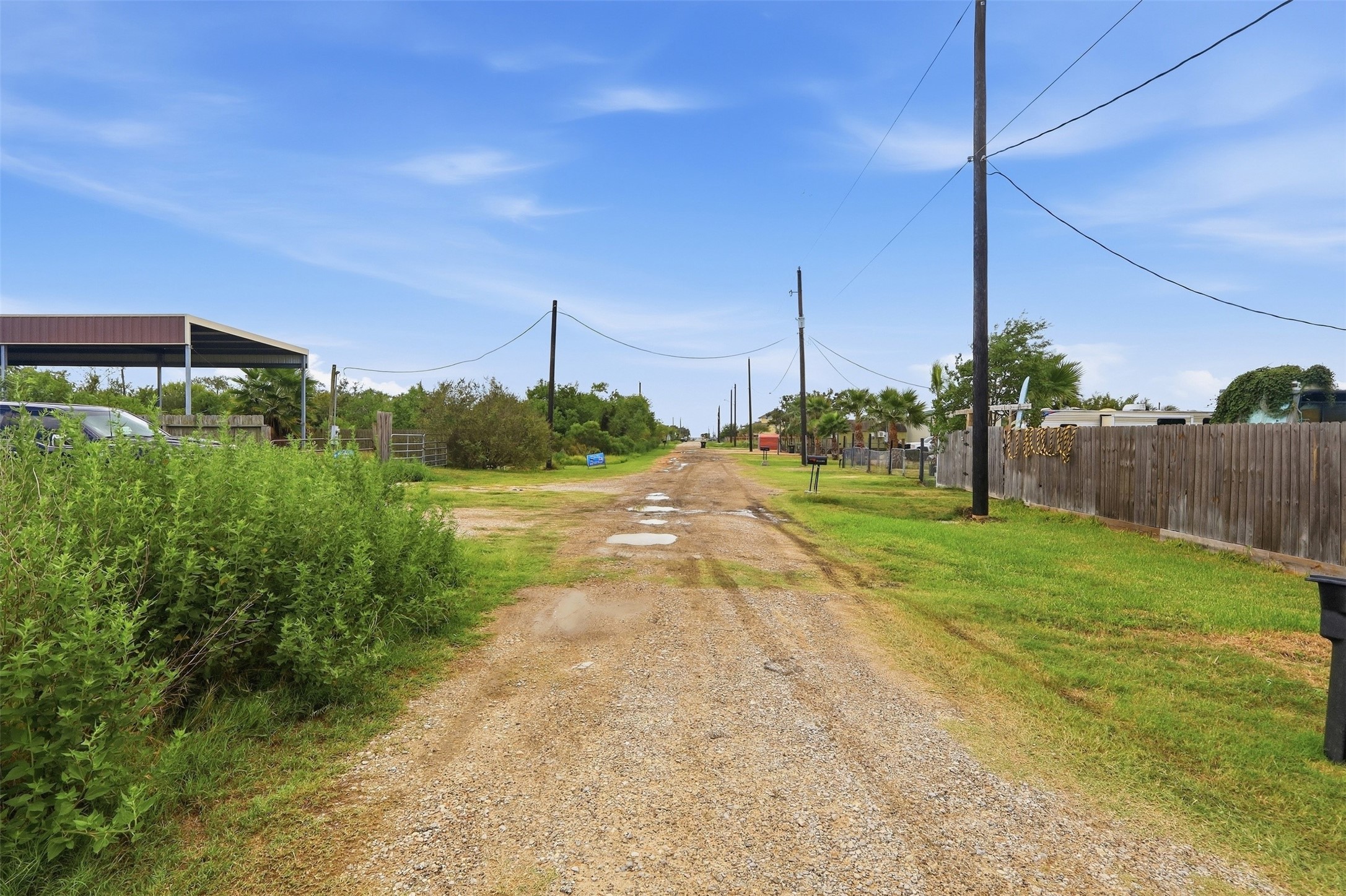 Gravel side road running along the property, with utility poles overhead. Neighboring lots are fenced and developed, and the street leads in the direction of the bay.