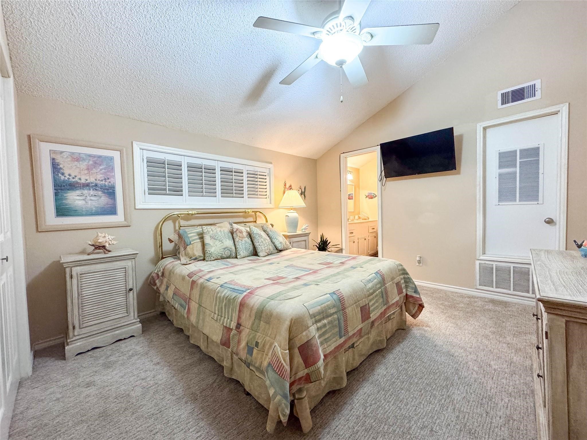 Cozy bedroom with soft lighting, featuring a comfortable bed, ceiling fan, and neutral decor. The room includes a wall-mounted TV, adjacent bathroom, and ample natural light from a window with shutters.