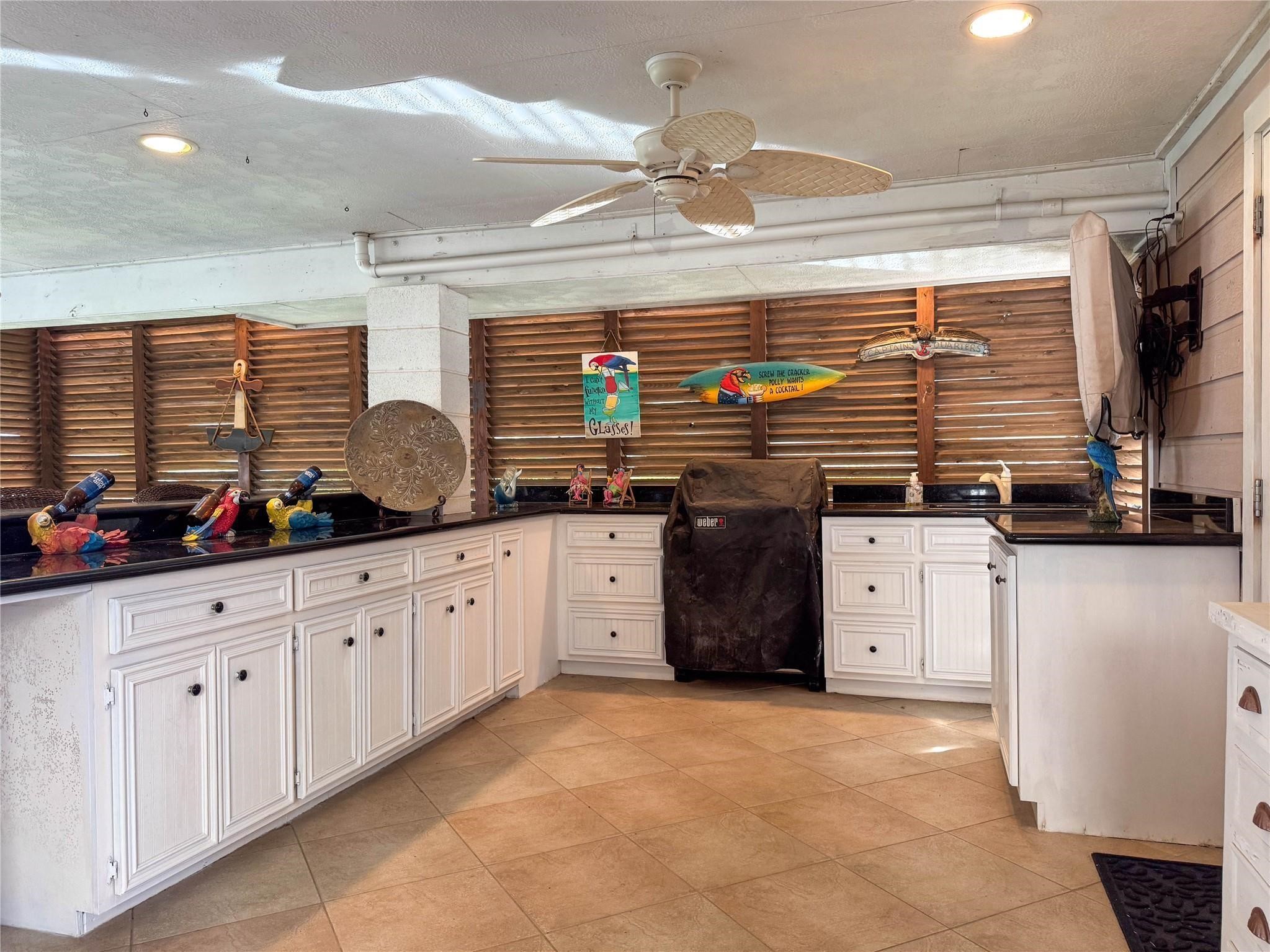 This photo showcases an inviting outdoor kitchen with ample counter space, white cabinetry, and decorative accents. The area includes a covered grill, ceiling fan, and wooden shutters, creating a relaxed, tropical atmosphere perfect for entertaining.