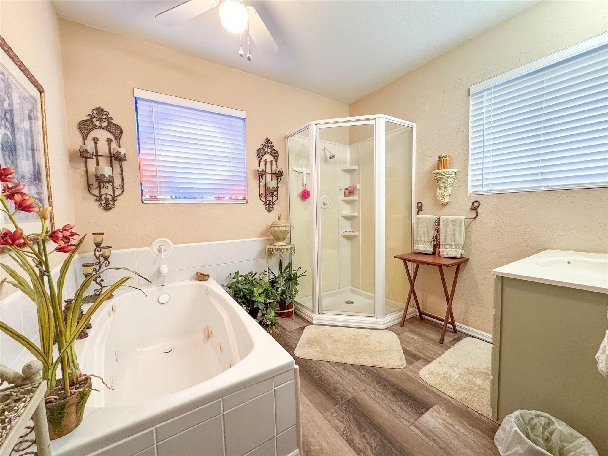This bathroom features a cozy layout with a corner shower, a spacious soaking tub, and tasteful decor. It has warm beige walls, wood-style flooring, and ample natural light from two windows, creating a relaxing atmosphere.