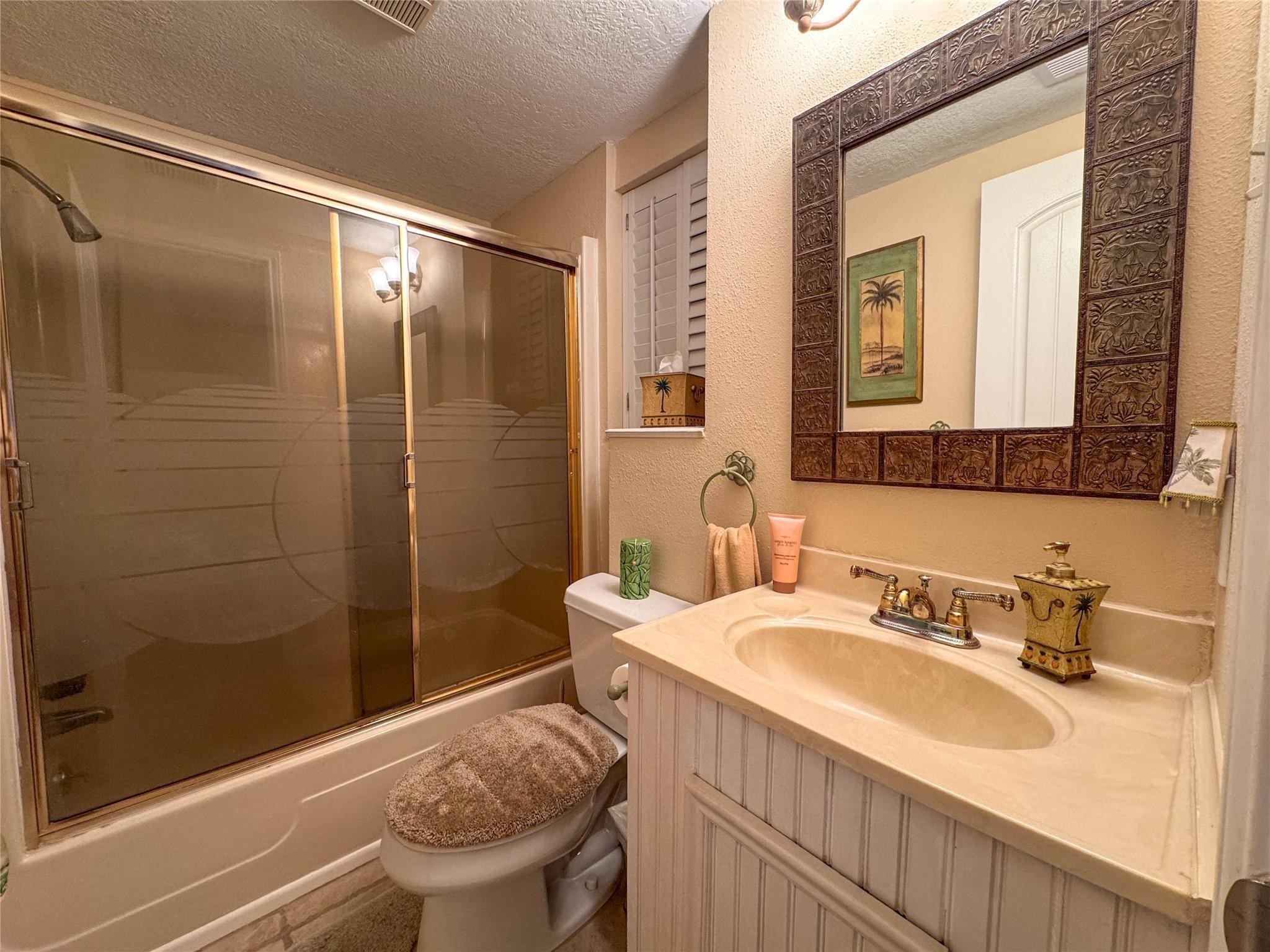 This cozy bathroom features a bathtub with sliding glass doors, a toilet with a plush cover, and a classic vanity with a decorative framed mirror. The warm lighting and palm-themed decor add a touch of elegance.