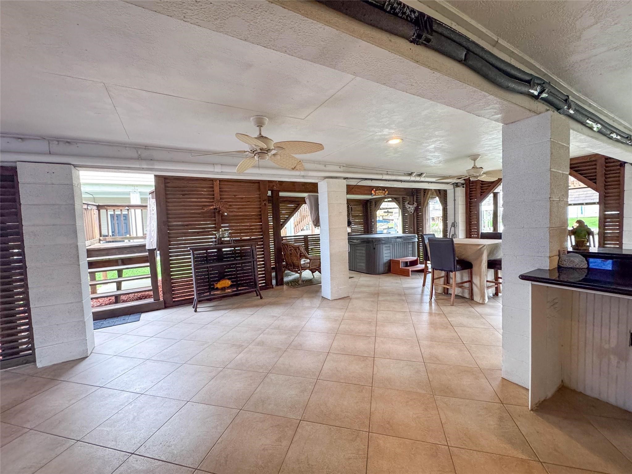This spacious covered patio features tiled flooring, ceiling fans, and ample seating areas, perfect for outdoor entertaining. The space includes a bar area and a hot tub, with wooden privacy screens providing a cozy and inviting atmosphere.