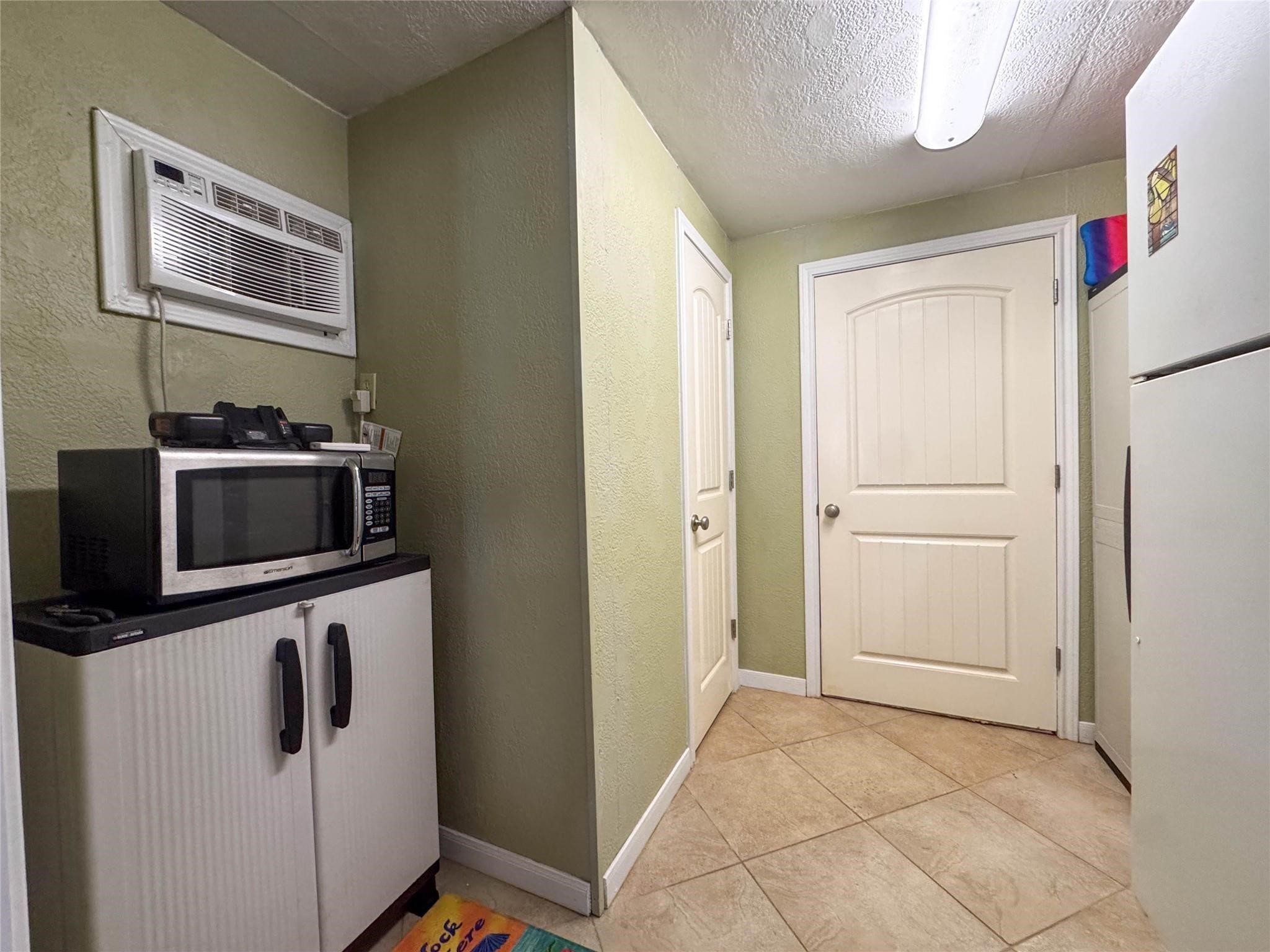 Compact utility space with tile flooring, featuring a wall-mounted air conditioner, a microwave atop a storage cabinet, and two white doors. Neutral wall color and ceiling lighting create a simple, functional area.