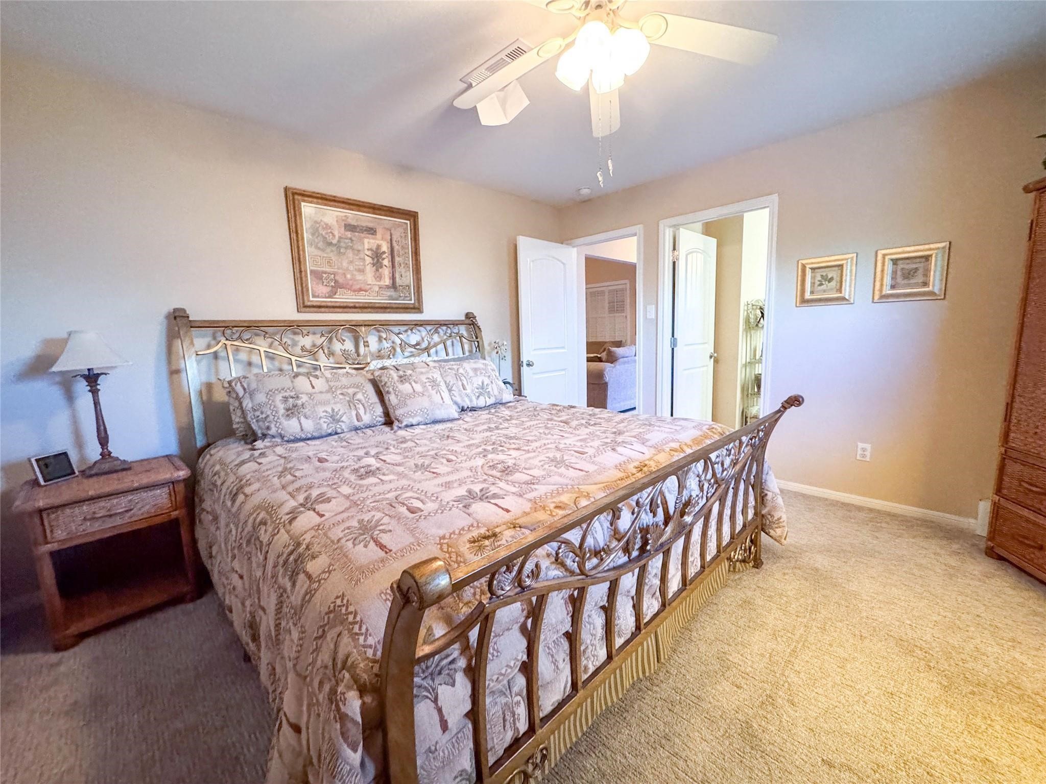 Cozy bedroom featuring a stylish metal bed frame, soft carpeting, and warm neutral walls. It includes a ceiling fan, wooden nightstand with a lamp, and tasteful wall art, creating a welcoming atmosphere.