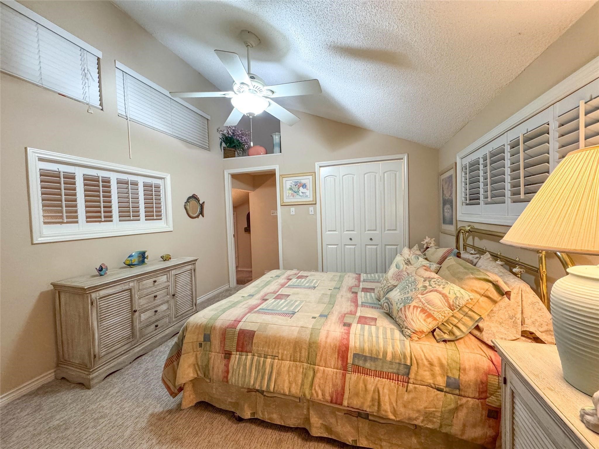 Cozy bedroom with vaulted ceilings, featuring a ceiling fan and soft carpeting. It includes a large bed with patterned bedding, ample closet space, and natural light from multiple windows with shutters, creating a warm and inviting atmosphere.