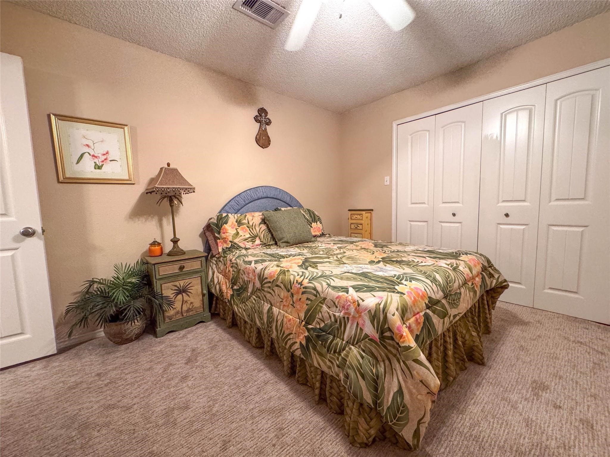Cozy bedroom with a tropical theme, featuring a floral bedspread, a ceiling fan, and ample closet space. Warm beige walls and carpet create a welcoming atmosphere.