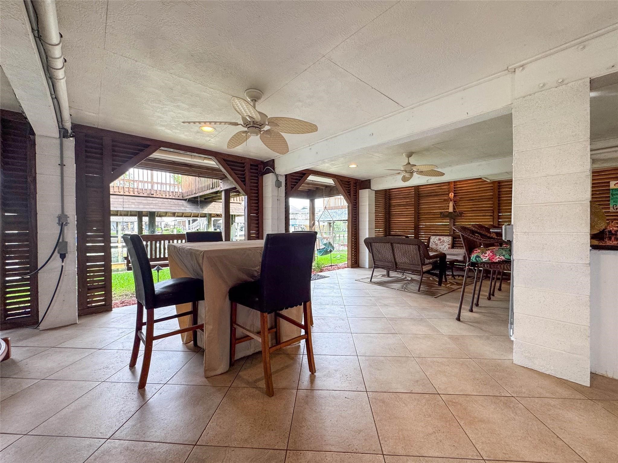This spacious covered patio features tiled flooring, ceiling fans, and ample seating areas, perfect for outdoor entertaining. It offers a cozy, open-air feel with wooden slat walls and a view of the surrounding greenery.