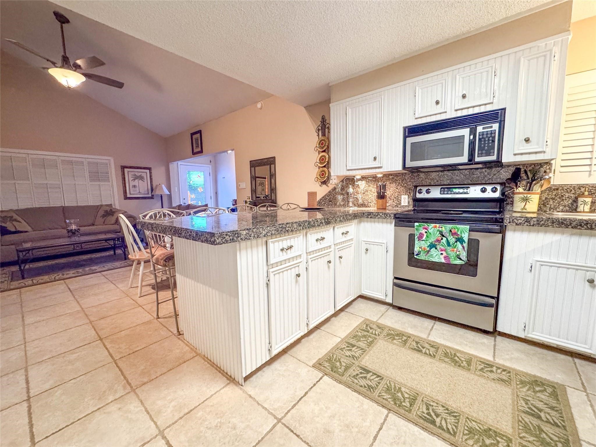 This inviting open-concept space features a cozy living area adjacent to a modern kitchen with granite countertops, white cabinetry, and stainless steel appliances. The tile flooring and vaulted ceiling enhance the room's spacious feel, perfect for relaxed living and entertaining.