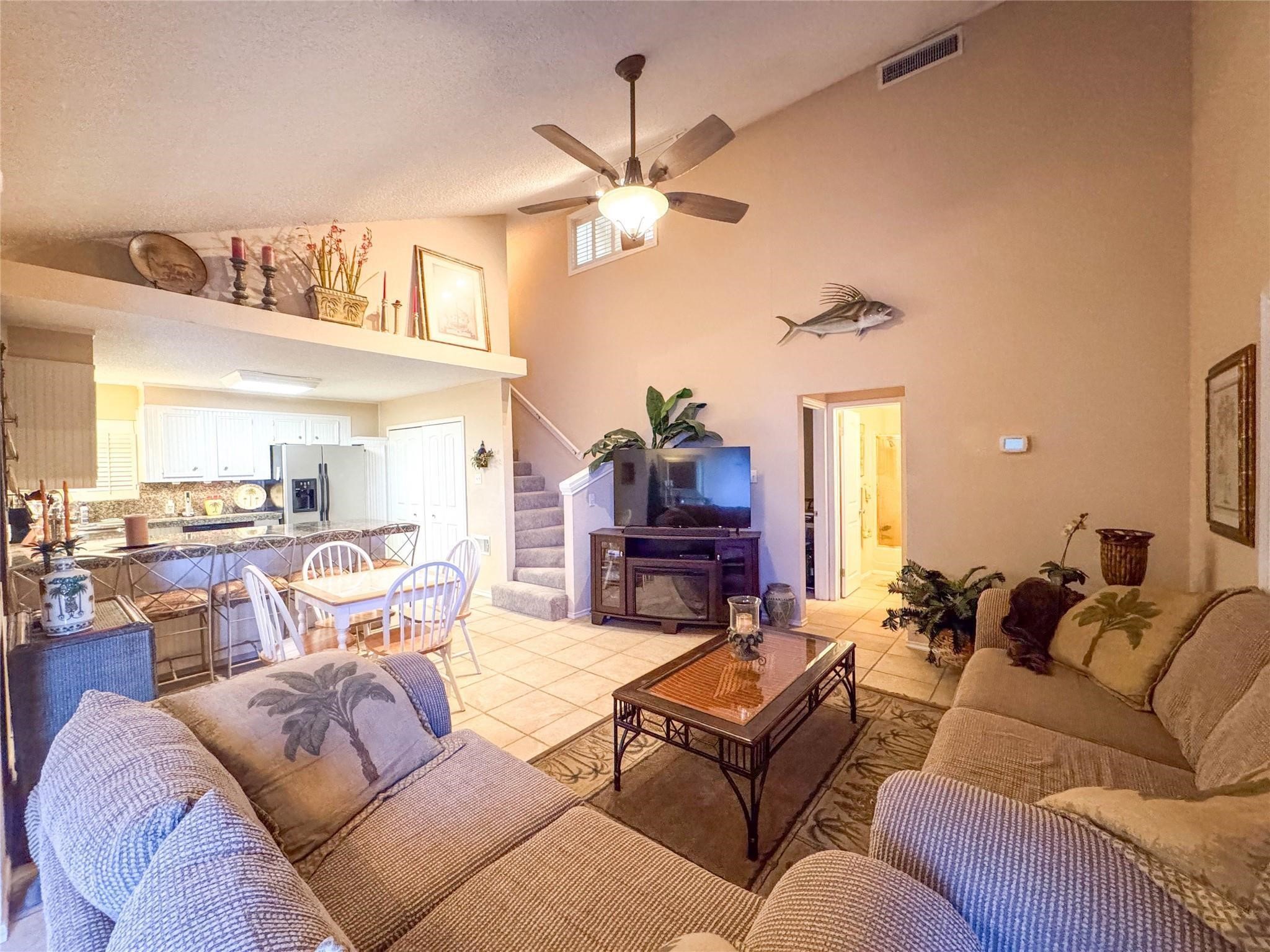 This inviting living space features high ceilings and an open layout with cozy seating, a ceiling fan, and decorative touches. The adjacent kitchen and dining area are bright and functional, with a staircase leading to the upper floor. Ideal for comfortable living and entertaining.
