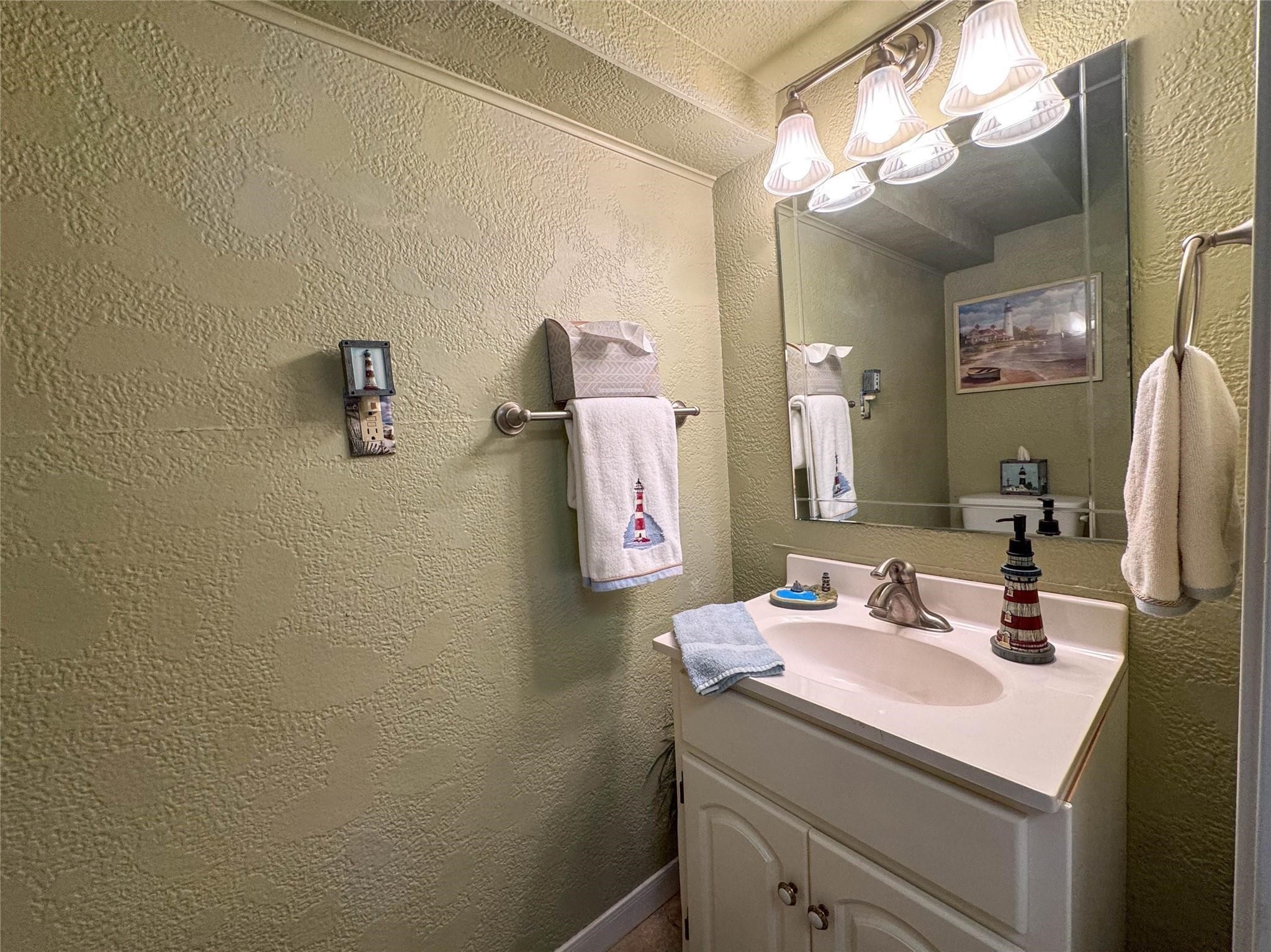 This bathroom features textured green walls, a large mirror with overhead lighting, and a white vanity. Nautical-themed accents add charm, including lighthouse decor and towels.