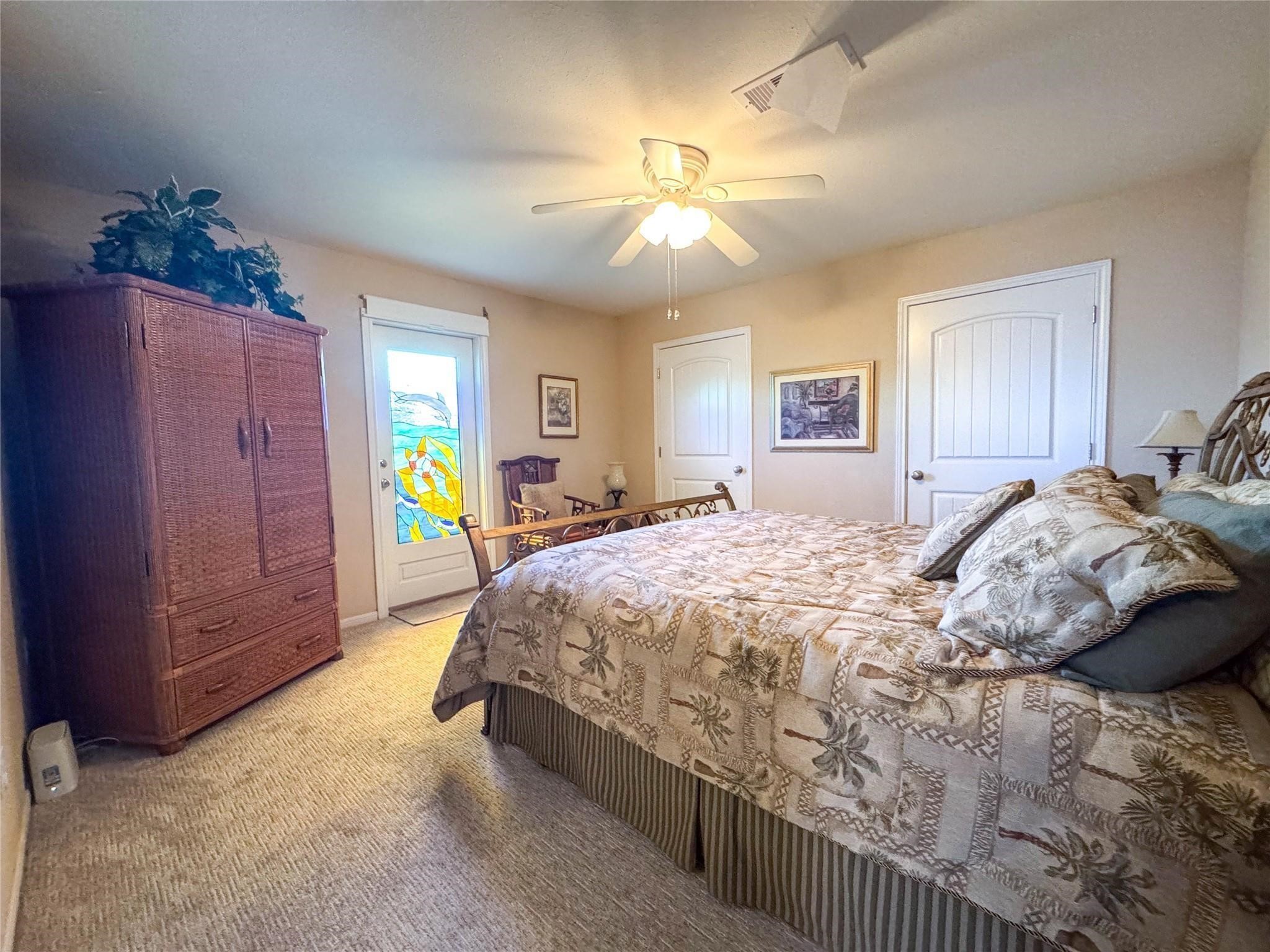 This cozy bedroom features a ceiling fan, a wicker armoire, and a bed with a tropical-themed comforter. A stained glass door adds a unique touch, complemented by neutral walls and carpet.