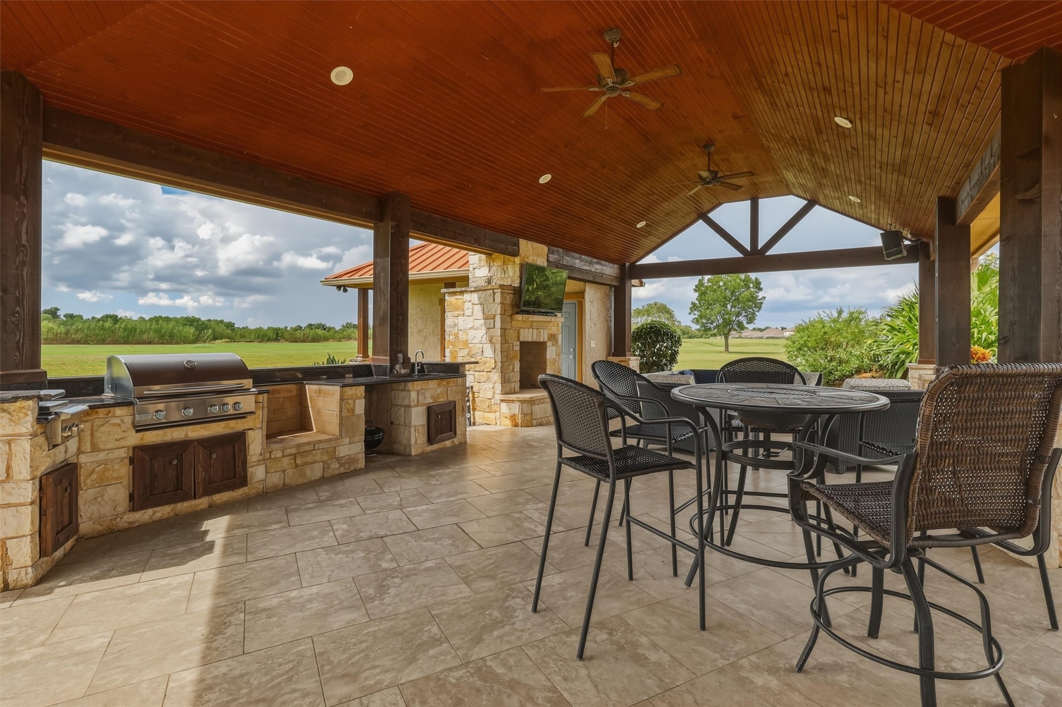 This impressive outdoor living space features a built-in grill station, stone fireplace, and wall-mounted TV, all beneath a soaring wood-paneled ceiling. Perfect for entertaining while enjoying views of the surrounding acreage.