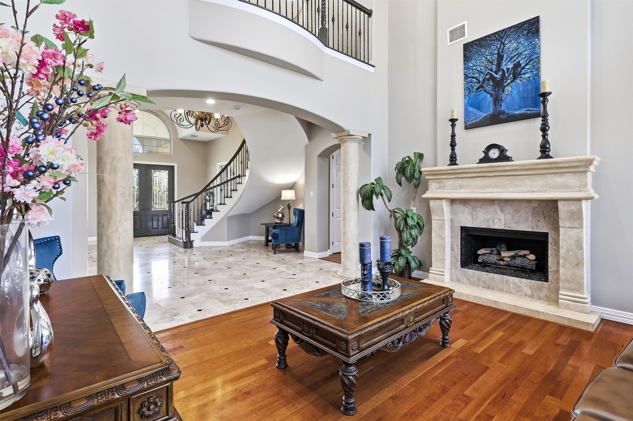This spacious living area showcases a striking stone fireplace with a custom mantel, soaring ceilings, and warm hardwood flooring. An open view to the curved staircase and entry beyond adds to the home's grand, connected layout.