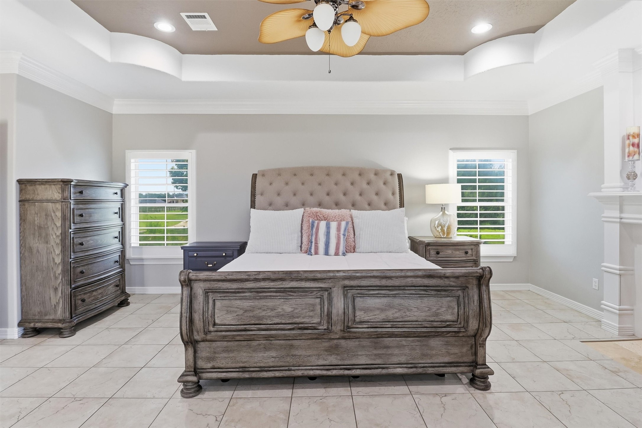 This luxurious primary bedroom offers a tray ceiling, a cozy fireplace, and abundant natural light from surrounding windows. Tile flooring and generous space for a sitting area make this a true private retreat.