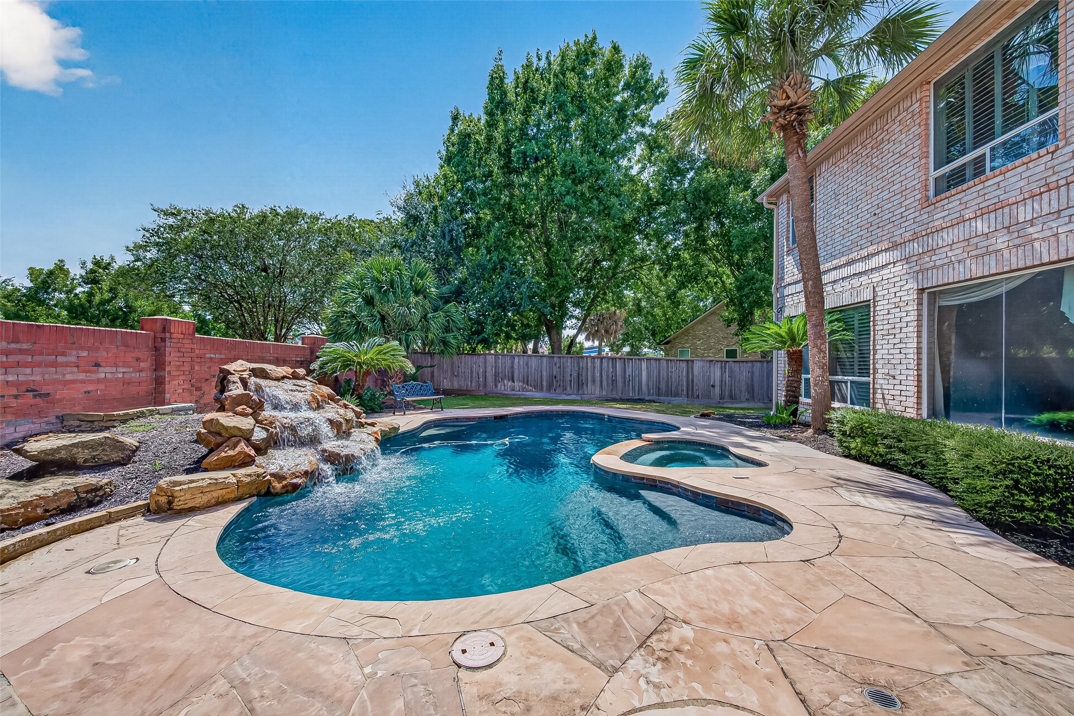 A stunning backyard oasis featuring a sparkling pool with a cascading waterfall, surrounded by lush landscaping and ample patio space for relaxing or entertaining.