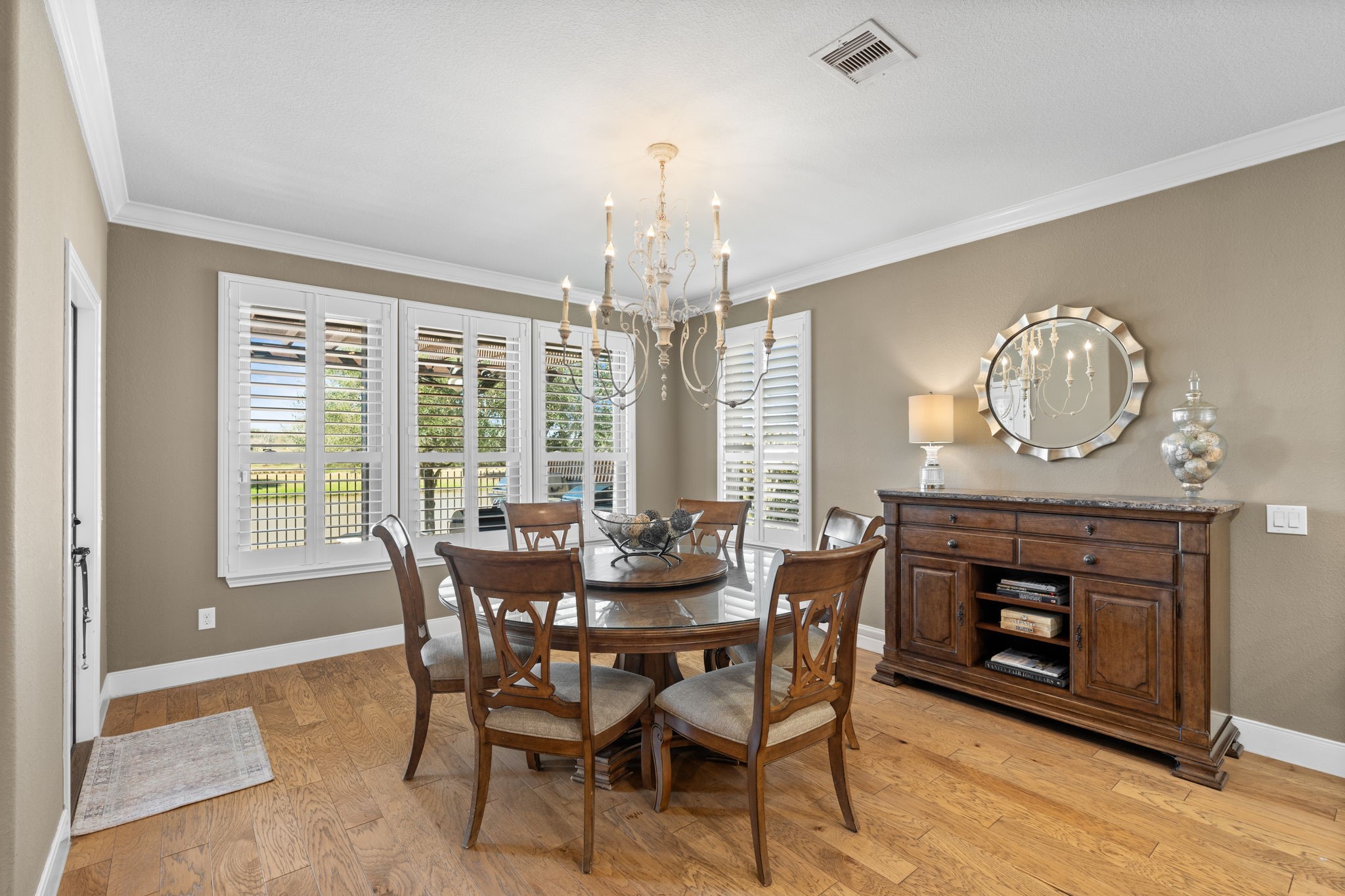 Spacious and elegantly appointed dining area with a modern hanging light fixture, hardwood floors, and neutral colors. White crown molding adds a classic touch, and the room flows seamlessly into adjacent living spaces