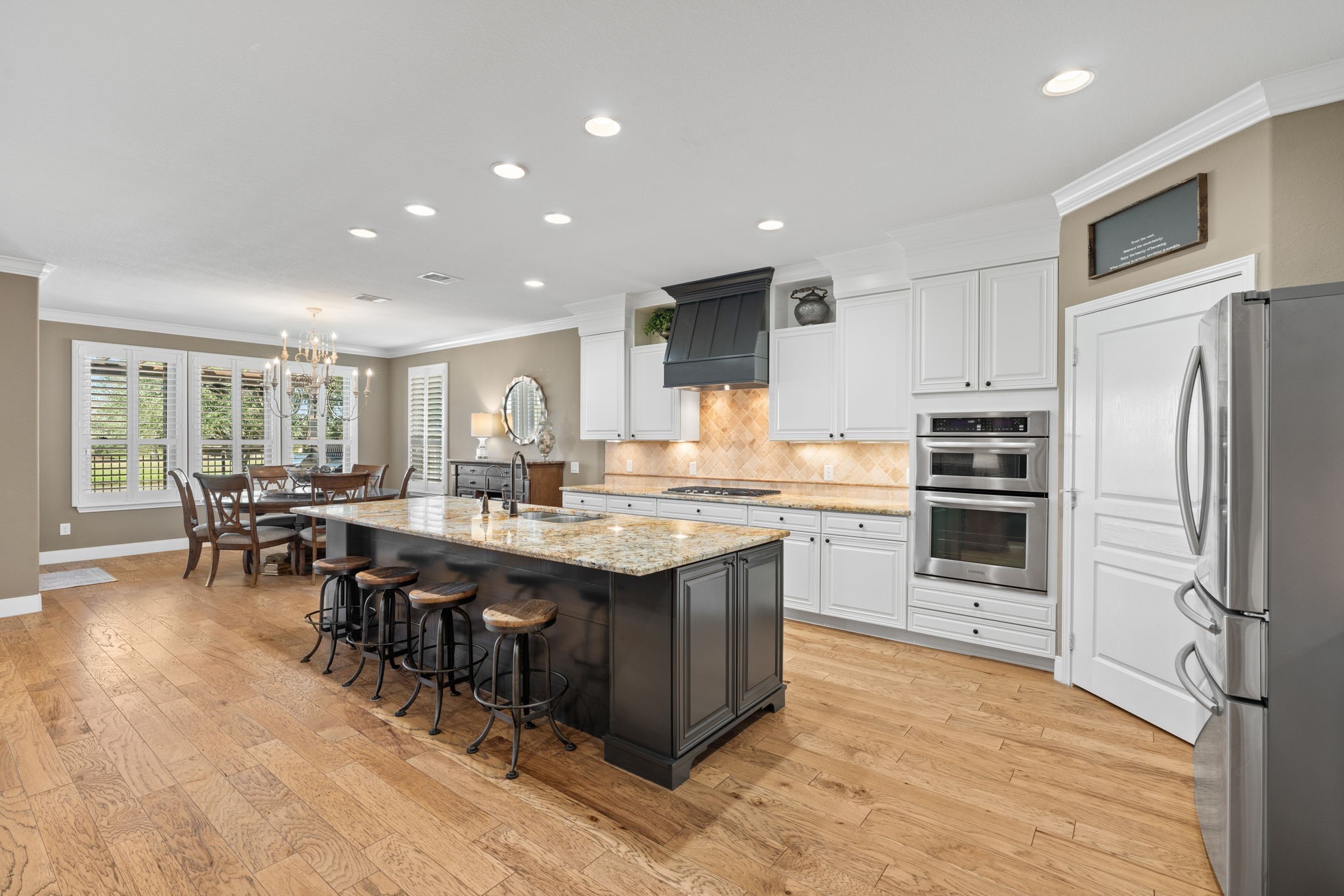 This is a spacious, modern kitchen with wooden cabinetry, granite countertops, and stainless-steel appliances. The open-plan layout includes a spacious island with exotic granite and plenty of recessed lights. The kitchen flows into a dining area, creating an inviting space for entertaining and daily living. Pull outs in bottom cabinets