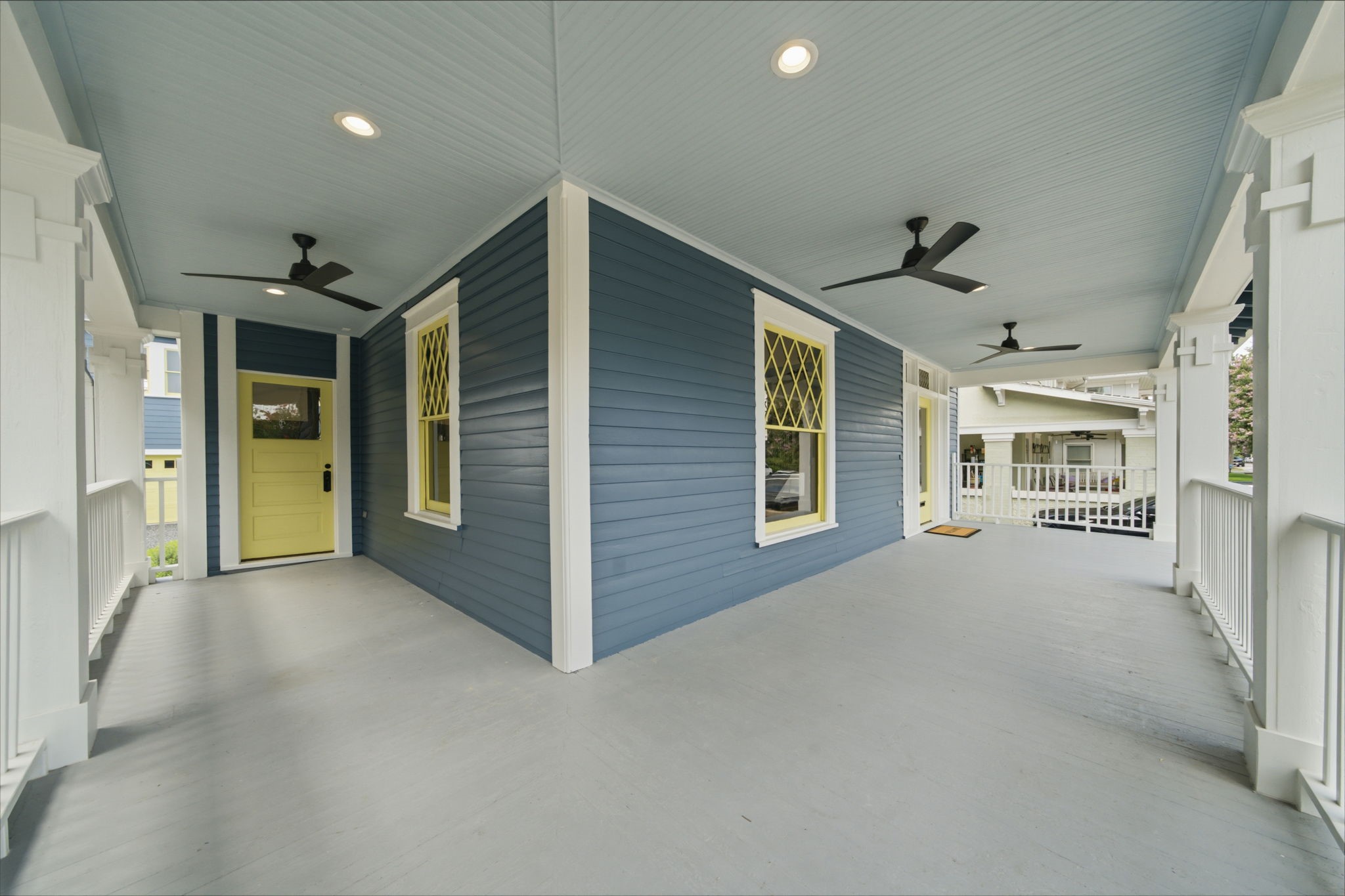 Charming wraparound porch with elegant blue siding, accented by a cheerful yellow door and stylish ceiling fans, perfect for relaxing and entertaining.