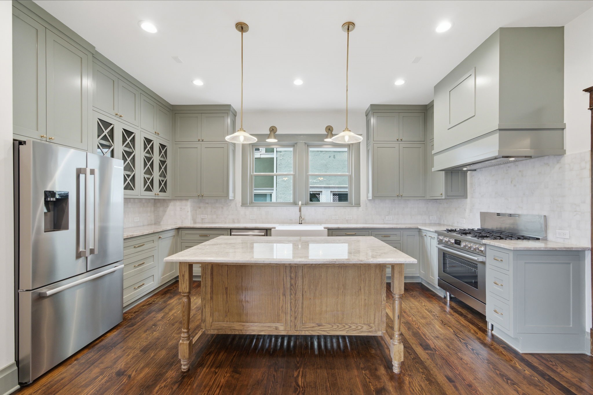 A spacious, modern kitchen boasts custom cabinetry, a large farm table inspired island with a beveled countertop, and high-end stainless steel appliances, complemented by warm wooden flooring and ample natural light.