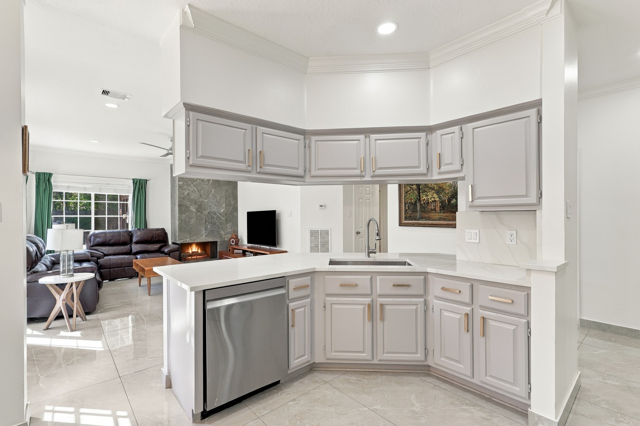 Thoughtfully designed kitchen features a wraparound breakfast bar with generous counter space, abundant gray cabinetry accented by brushed gold hardware, and a stainless steel dishwasher. The open pass-through maintains a connection to the living area, while crown molding, recessed lighting, and polished flooring complete the space.