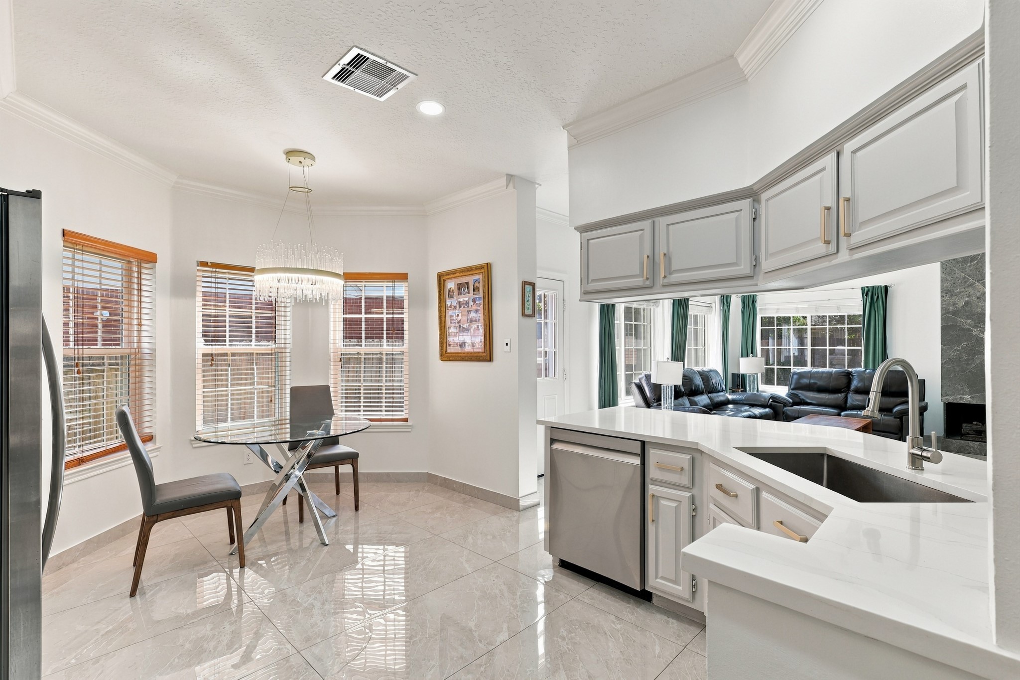 The kitchen opens effortlessly to a sunlit breakfast nook surrounded by windows, creating a welcoming space for casual dining. A convenient pass-through overlooks the living room, while white countertops, gray cabinetry with gold hardware, stainless steel dishwasher, and polished flooring tie the spaces together beautifully.