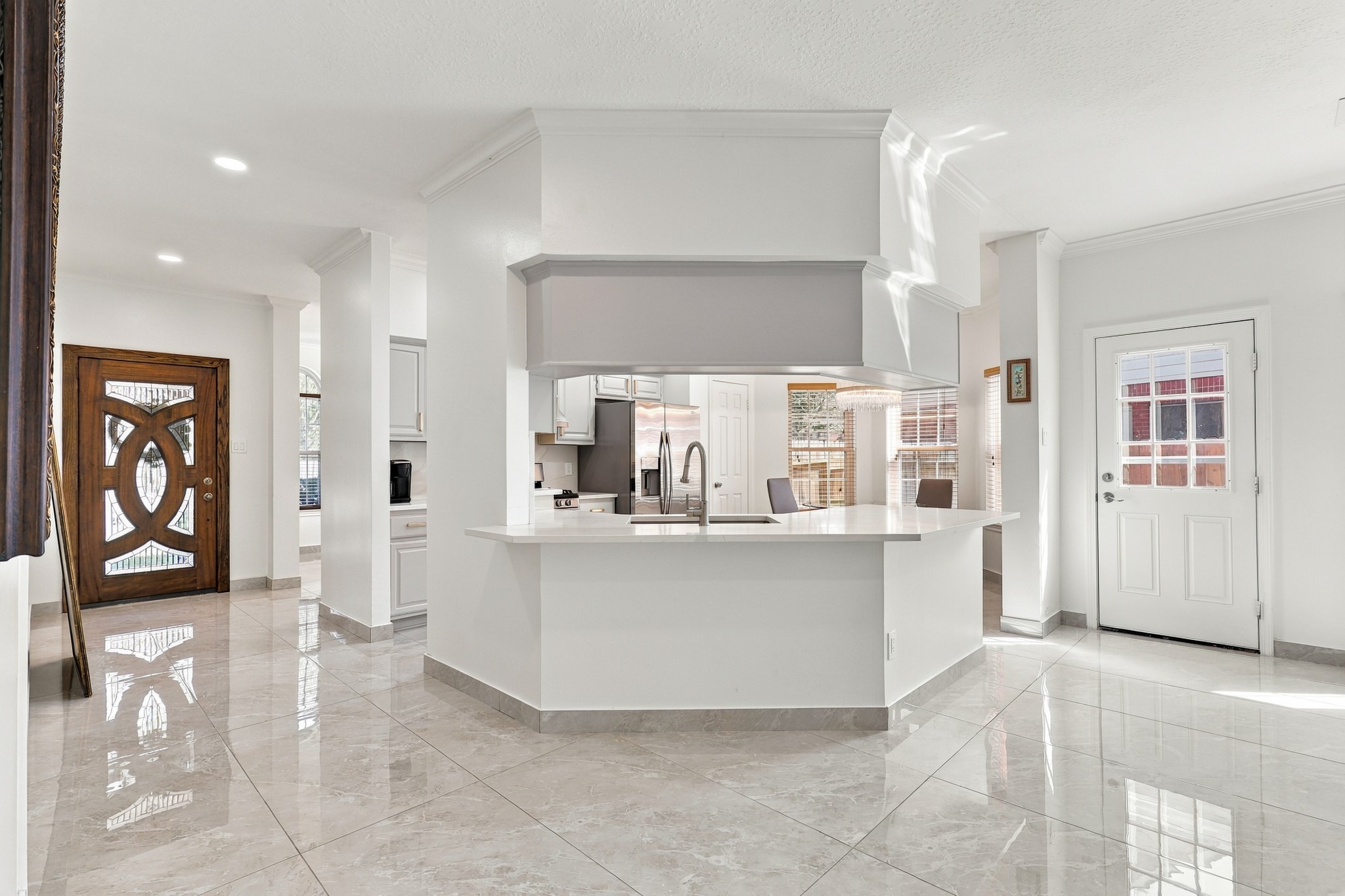 Bright, open-concept layout featuring a spacious kitchen with wraparound breakfast bar, crisp white countertops, and views into the adjoining living and dining areas. Soaring ceilings, decorative columns, crown molding, recessed lighting, and polished large-format tile flooring enhance the home's airy feel, while the distinctive wood entry door adds architectural character. Bright, open-concept layout featuring a spacious kitchen with wraparound breakfast bar, crisp white countertops, and views into the adjoining living and dining areas. Soaring ceilings, decorative columns, crown molding, recessed lighting, and polished large-format tile flooring enhance the home's airy feel, while the distinctive wood entry door adds architectural character.