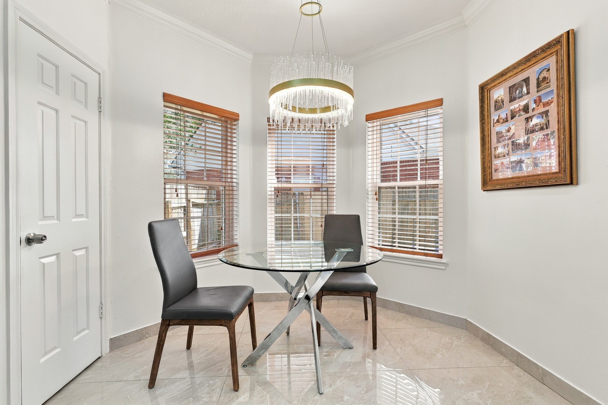 Charming breakfast nook featuring a curved bay of three windows that fills the space with natural light. An elegant crystal-style chandelier with gold accents adds a sophisticated touch, while crown molding, wood-trimmed blinds, and polished marble tile flooring complete this inviting dining space.