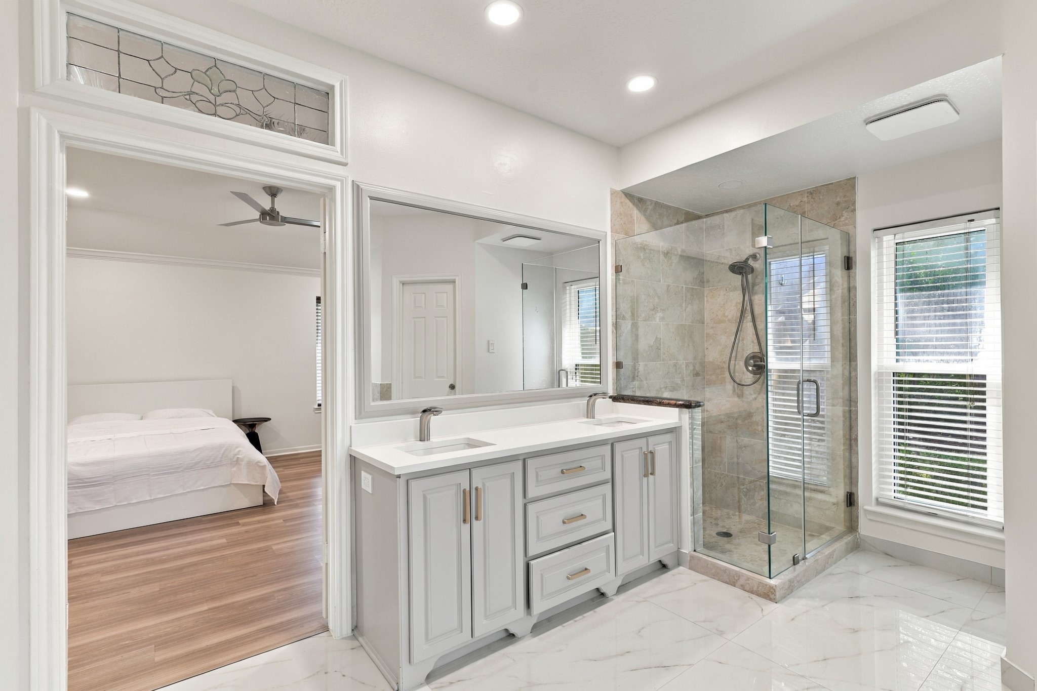 Beautifully appointed primary bathroom featuring a double-sink vanity with generous cabinetry, sleek countertops, and brushed gold hardware. A spacious glass-enclosed shower with floor-to-ceiling tile, elegant marble flooring, and natural light from the large window complete the space.