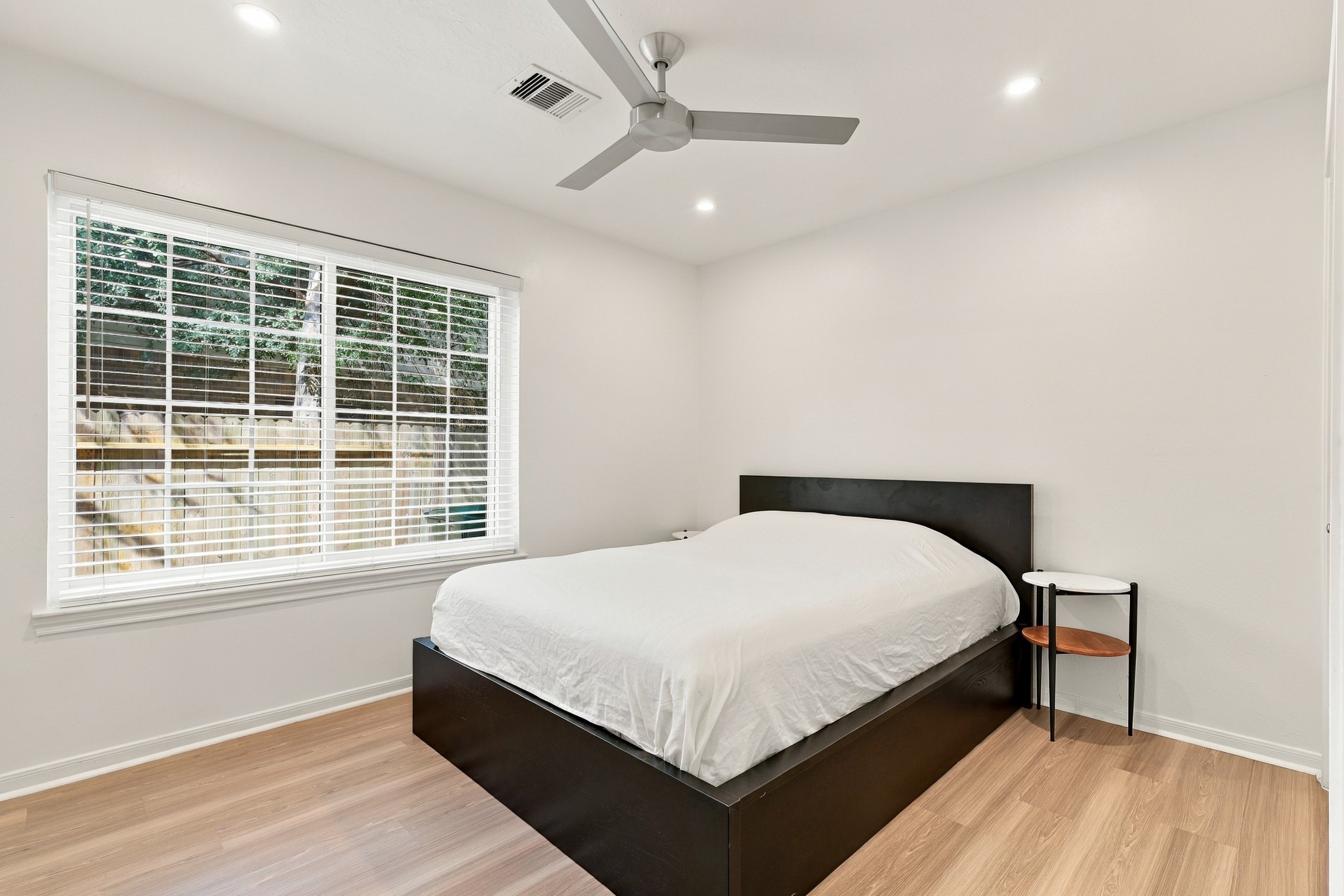 Spacious bedroom featuring warm wood flooring, a large window overlooking the backyard, recessed lighting, and a modern ceiling fan. Crisp white walls and abundant natural light create a bright, inviting retreat.