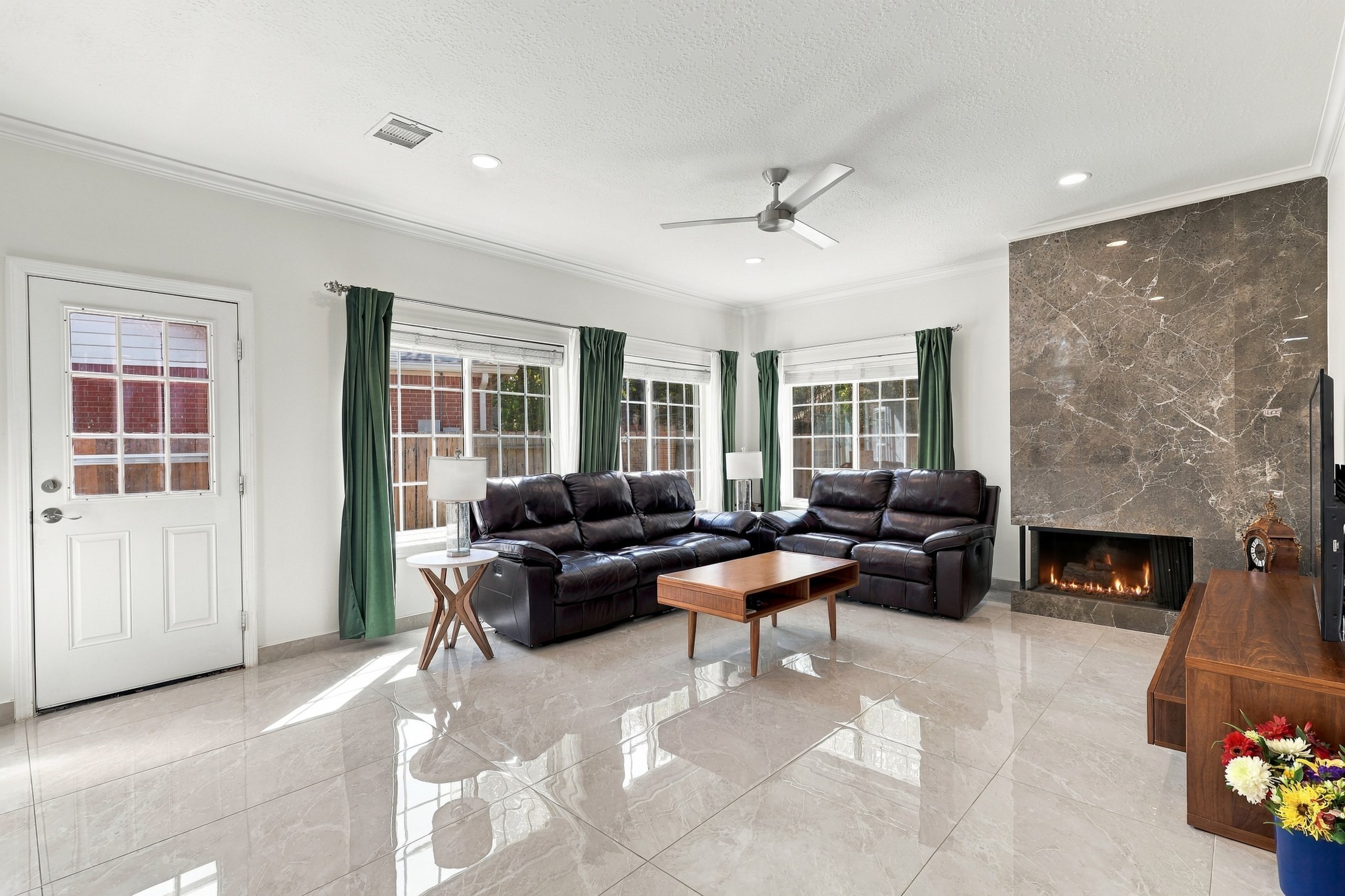 Spacious living area filled with natural light from multiple large windows, featuring glossy marble flooring, crown molding, recessed lighting, and a modern ceiling fan. A striking floor-to-ceiling, marble fireplace surround creates a dramatic focal point, while the open layout offers plenty of room for comfortable seating and entertaining. Spacious living area filled with natural light from multiple large windows, featuring glossy marble flooring, crown molding, recessed lighting, and a modern ceiling fan. A striking floor-to-ceiling, marble fireplace surround creates a dramatic focal point, while the open layout offers plenty of room for comfortable seating and entertaining.