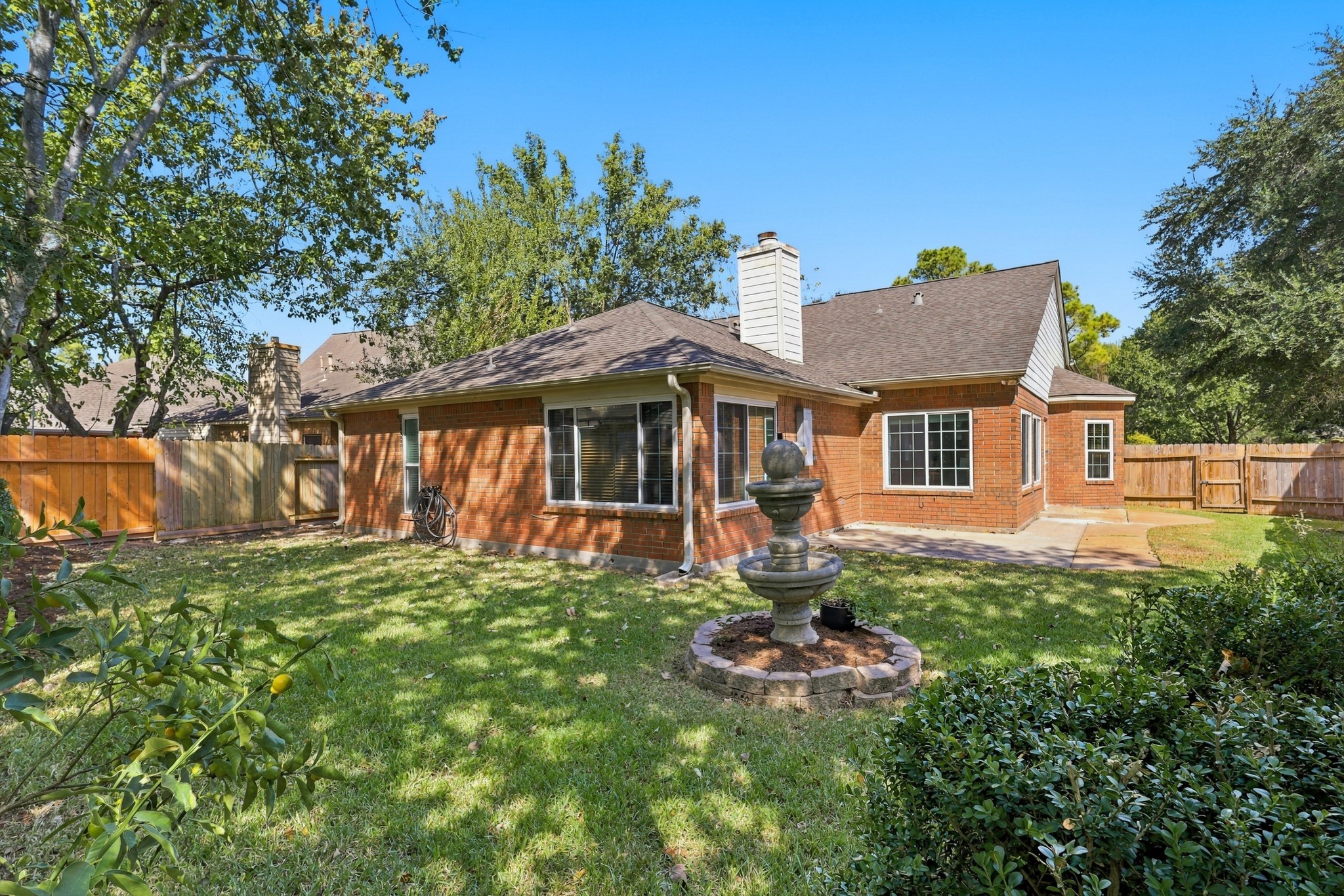 The spacious backyard features a classic brick exterior, mature shade trees, lush greenery, and a fully fenced lawn. A decorative tiered fountain creates a charming focal point, while the generous concrete patio provides an ideal space for outdoor dining, entertaining, or relaxing. Large windows overlook the backyard, bringing natural light into the home and connecting the indoor and outdoor living spaces.